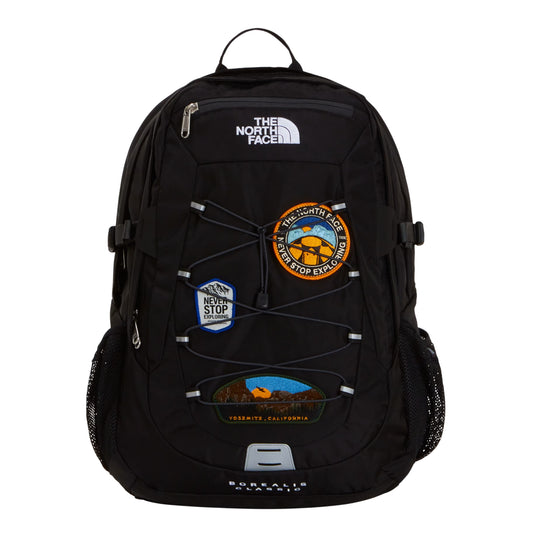 Mochila The North Face Borealis Classic Patch em TNF Black — vista frontal, produto sem modelo, 3 patches bordados: circular laranja/amarelo "Never Stop Exploring", hexagonal azul com montanhas e oval Yosemite California, sistema de cordas elásticas pretas, logo TNF bordado em branco e bolsos laterais em malha preta.