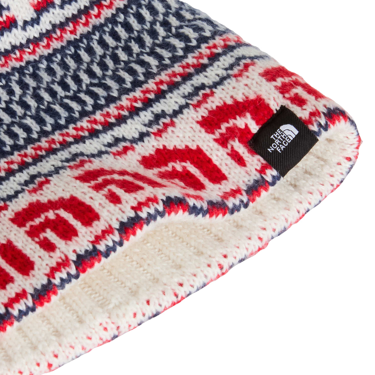 Pormenor da malha jacquard do gorro The North Face Cabin Ski Tuke White Dune, com logótipos vermelhos e etiqueta preta na extremidade.