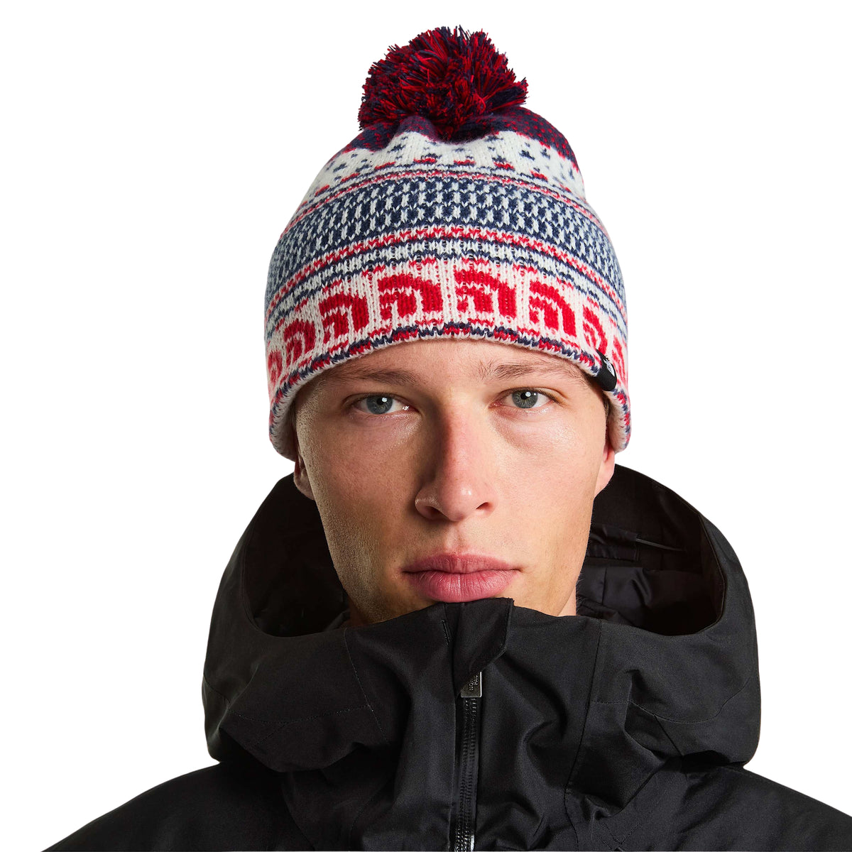 Modelo de frente com gorro The North Face Cabin Ski Tuke White Dune Jacquard, com pompom e padrão retro, e casaco preto.