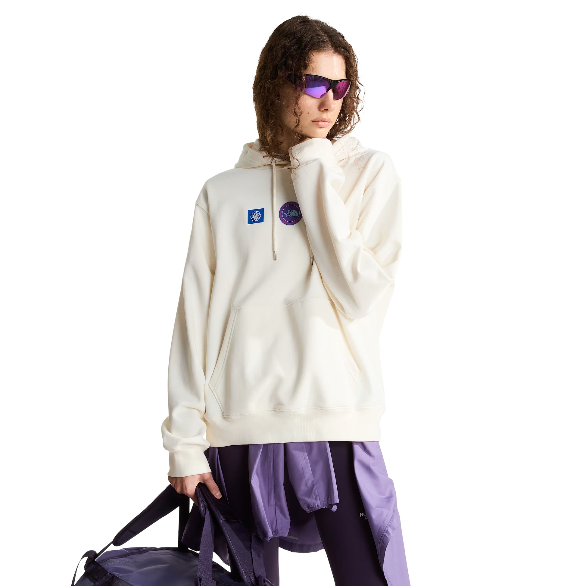Hoodie unissexo The North Face Explorer Badges Relaxed em White Dune - modelo feminino de frente a 3/4, cabelo encaracolado castanho, óculos de sol wrap roxos, capuz levantado com cordão branco, dois emblemas Explorer visíveis no peito (azul quadrado com globo e circular roxo "The North Face"), bolso canguru com mão dentro, bainha canelada e corte relaxado com ombros descaídos visíveis, calças TNF roxas, saco duffel TNF preto na mão direita, French Terry White Dune 360 g/m², fundo branco.