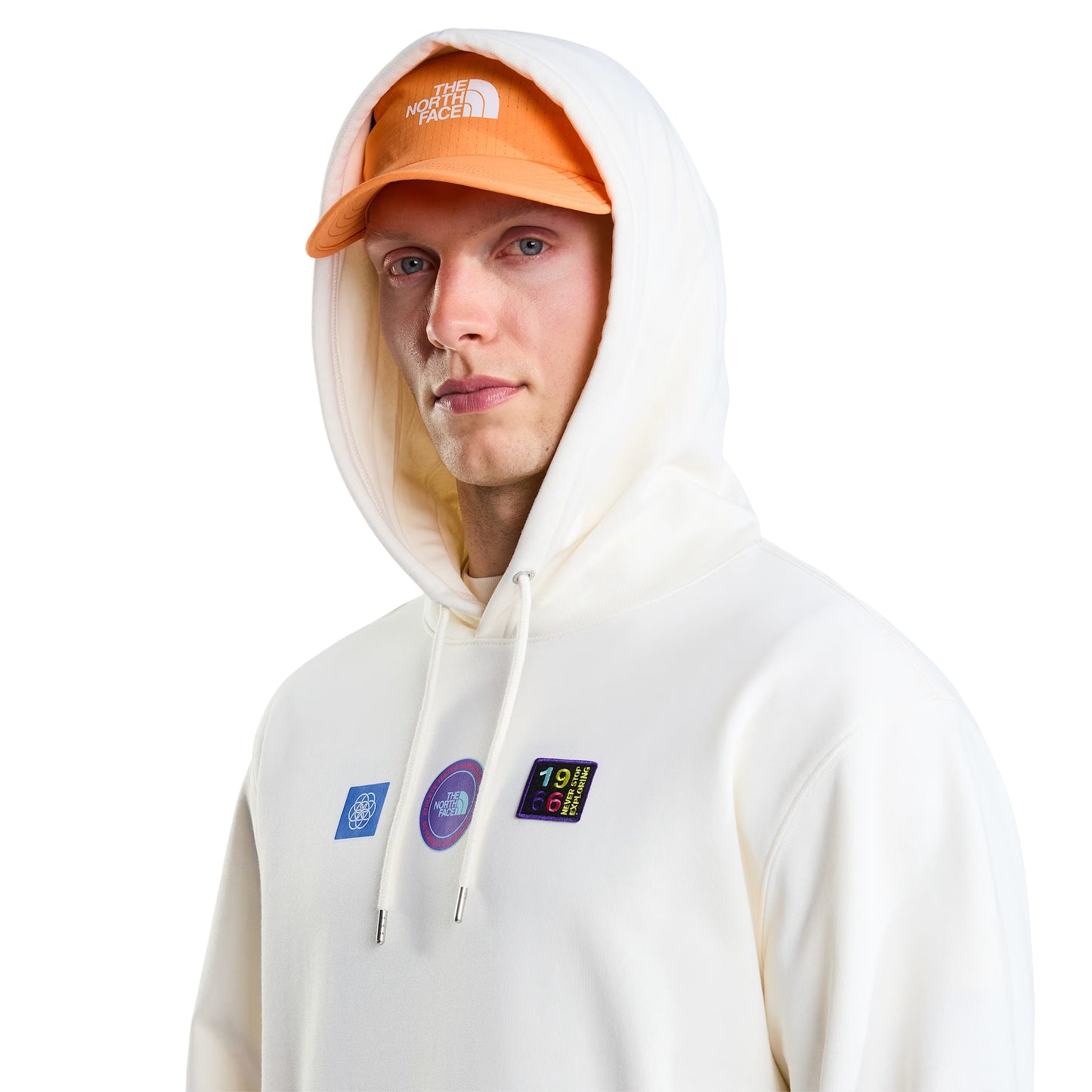 Hoodie unissexo The North Face Explorer Badges Relaxed em White Dune — modelo masculino de frente, meio corpo, boné técnico laranja TNF, óculos de sol wrap escuros, capuz levantado com cordão branco, detalhe dos três emblemas Explorer estampados no peito em close-up, French Terry White Dune 360 g/m², fundo branco.