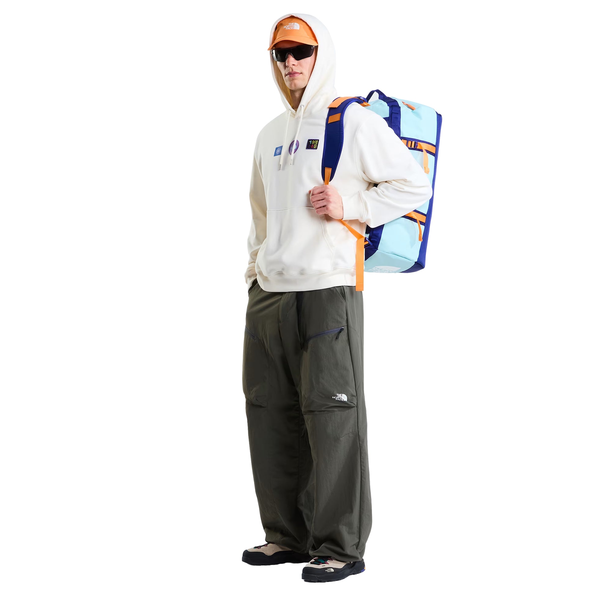 Hoodie unissexo The North Face Explorer Badges Relaxed em White Dune — modelo masculino de frente, corpo inteiro, boné técnico laranja TNF, óculos de sol wrap escuros, calças cargo verde-oliva TNF, sapatilhas de trail, mochila duffel TNF azul-claro e azul-marinho ao ombro, três emblemas Explorer estampados no peito visíveis, capuz levantado, bolso canguru e bainha canelada visíveis, fundo branco.
