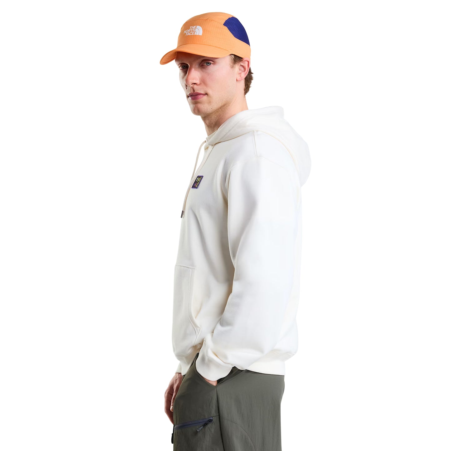 Hoodie unissexo The North Face Explorer Badges Relaxed em White Dune — modelo masculino de frente a 3/4, boné técnico laranja TNF, óculos de sol wrap escuros, capuz levantado com cordão branco, três emblemas Explorer estampados no peito visíveis, bolso canguru e bainha canelada visíveis, French Terry White Dune 360 g/m², fundo branco.