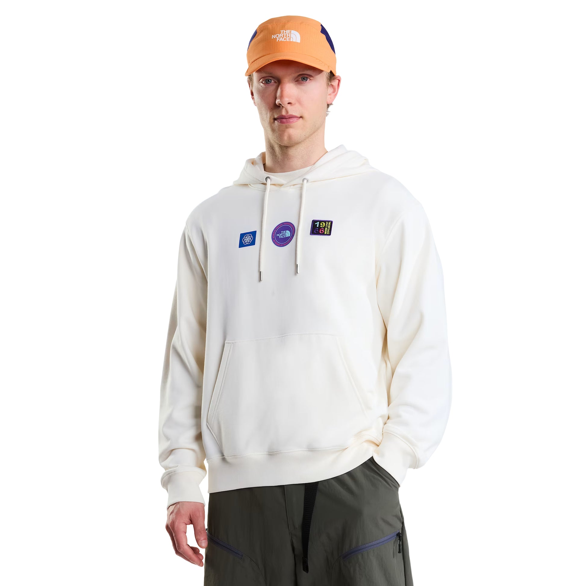 Hoodie unissexo The North Face Explorer Badges Relaxed em White Dune — modelo masculino de perfil esquerdo, boné técnico laranja TNF, óculos de sol wrap escuros, calças cargo verde-oliva, mochila duffel TNF azul-claro e azul-marinho às costas, capuz levantado, emblema "1966 Never Stop Exploring" parcialmente visível no peito, bolso canguru e bainha canelada visíveis, French Terry White Dune 360 g/m², fundo branco.