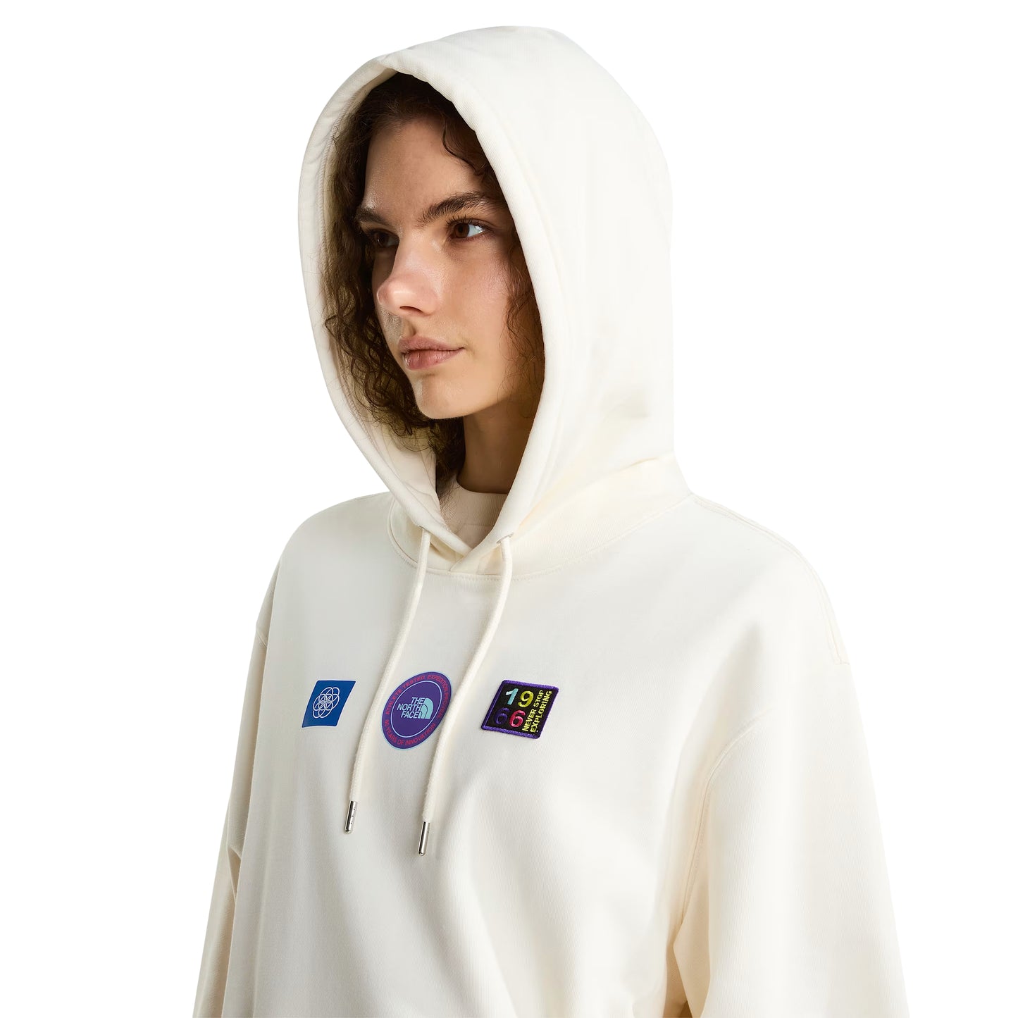 Hoodie unissexo The North Face Explorer Badges Relaxed em White Dune — modelo feminino de frente, meio corpo, cabelo encaracolado castanho, capuz levantado com cordão branco, detalhe dos três emblemas Explorer estampados em serigrafia à base de água no peito em close-up (azul quadrado com globo, circular roxo "The North Face", retangular escuro "1966 Never Stop Exploring" multicolor), French Terry White Dune 360 g/m², fundo branco.