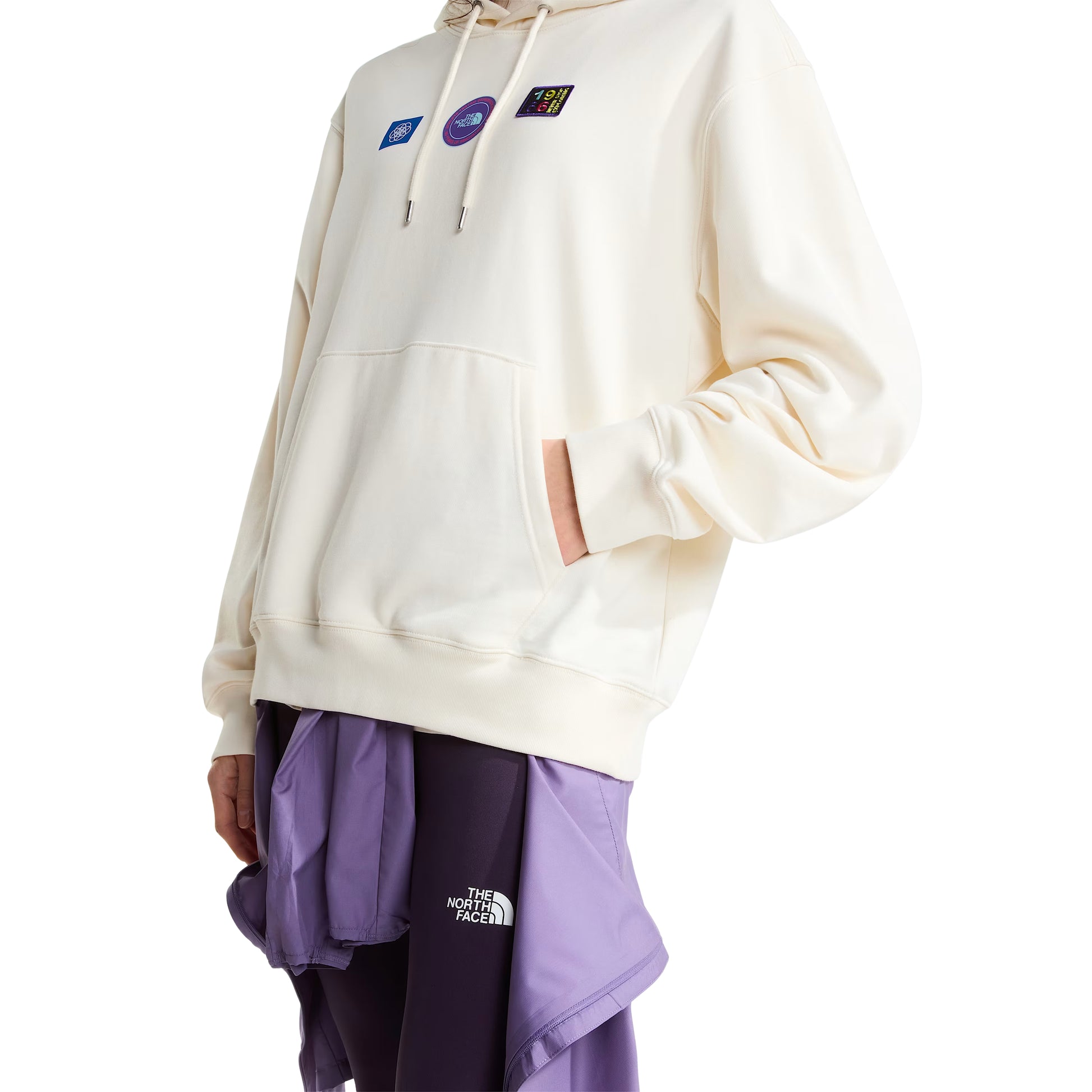 Hoodie unissexo The North Face Explorer Badges Relaxed em White Dune — modelo feminino de frente a 3/4, meio corpo, capuz levantado com cordão branco e ponteiras metálicas visíveis, três emblemas Explorer estampados em serigrafia à base de água no peito em close-up (azul quadrado com globo, circular roxo "The North Face", retangular escuro "1966 Never Stop Exploring" multicolor), bolso canguru com mão dentro, bainha canelada visível, calças TNF roxas e lilás sobrepostas com logótipo "The North Face" branco.