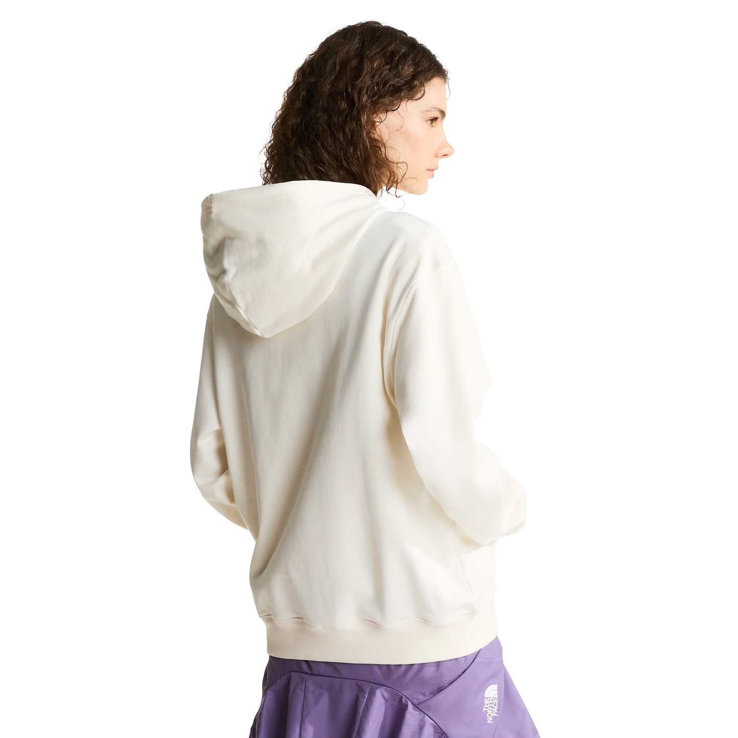 Hoodie unissexo The North Face Explorer Badges Relaxed em White Dune — modelo feminino de costas, cabelo encaracolado castanho, calças TNF roxas e lilás sobrepostas, capuz levantado, logótipo "The North Face" half-dome estampado em serigrafia à base de água no ombro direito nas costas visível, corte relaxado com ombros descaídos, French Terry White Dune 360 g/m², fundo branco.