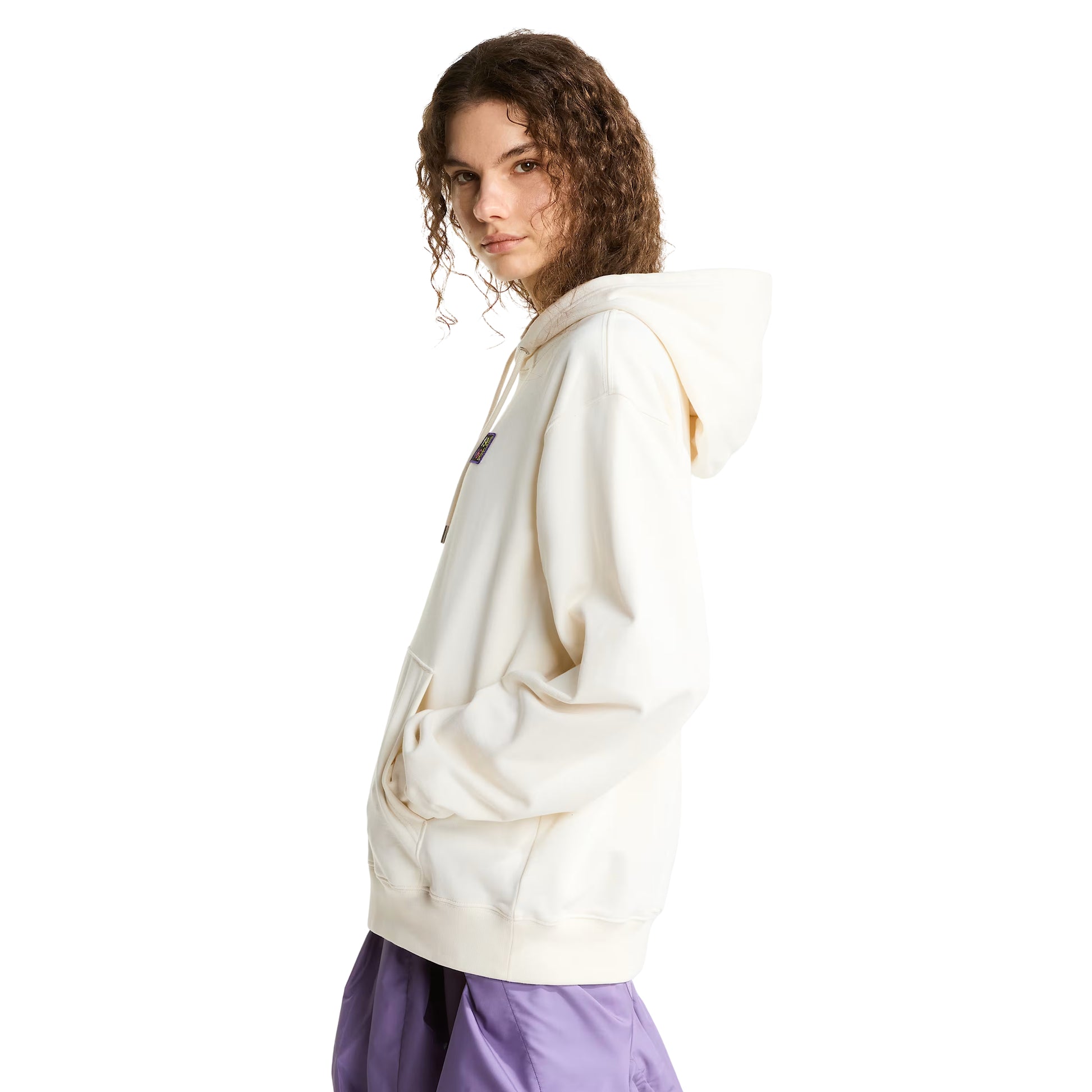 Hoodie unissexo The North Face Explorer Badges Relaxed em White Dune — modelo feminino de perfil direito, cabelo encaracolado castanho, capuz levantado com cordão laranja visível, emblema retangular escuro "1966 Never Stop Exploring" parcialmente visível no peito, mãos no bolso canguru, bainha canelada e corte relaxado com ombros descaídos visíveis, calças TNF roxas, French Terry White Dune 360 g/m², fundo branco.