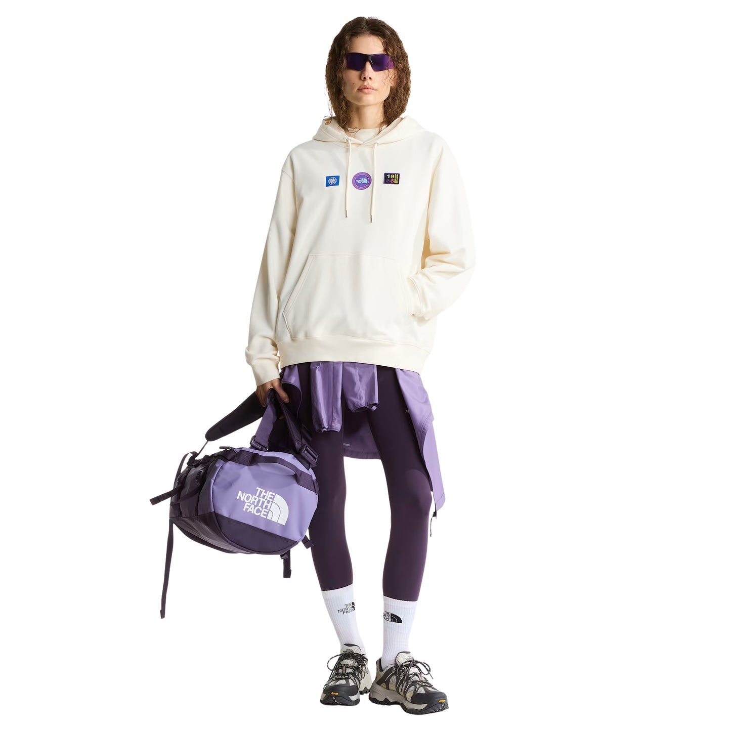 Hoodie unissexo The North Face Explorer Badges Relaxed em White Dune — modelo feminino de frente, corpo inteiro, calças TNF roxas e lilás sobrepostas, sapatilhas de trail, três emblemas Explorer estampados no peito visíveis, capuz levantado com cordão branco, bolso canguru e bainha canelada visíveis, corte relaxado com ombros descaídos, fundo branco.