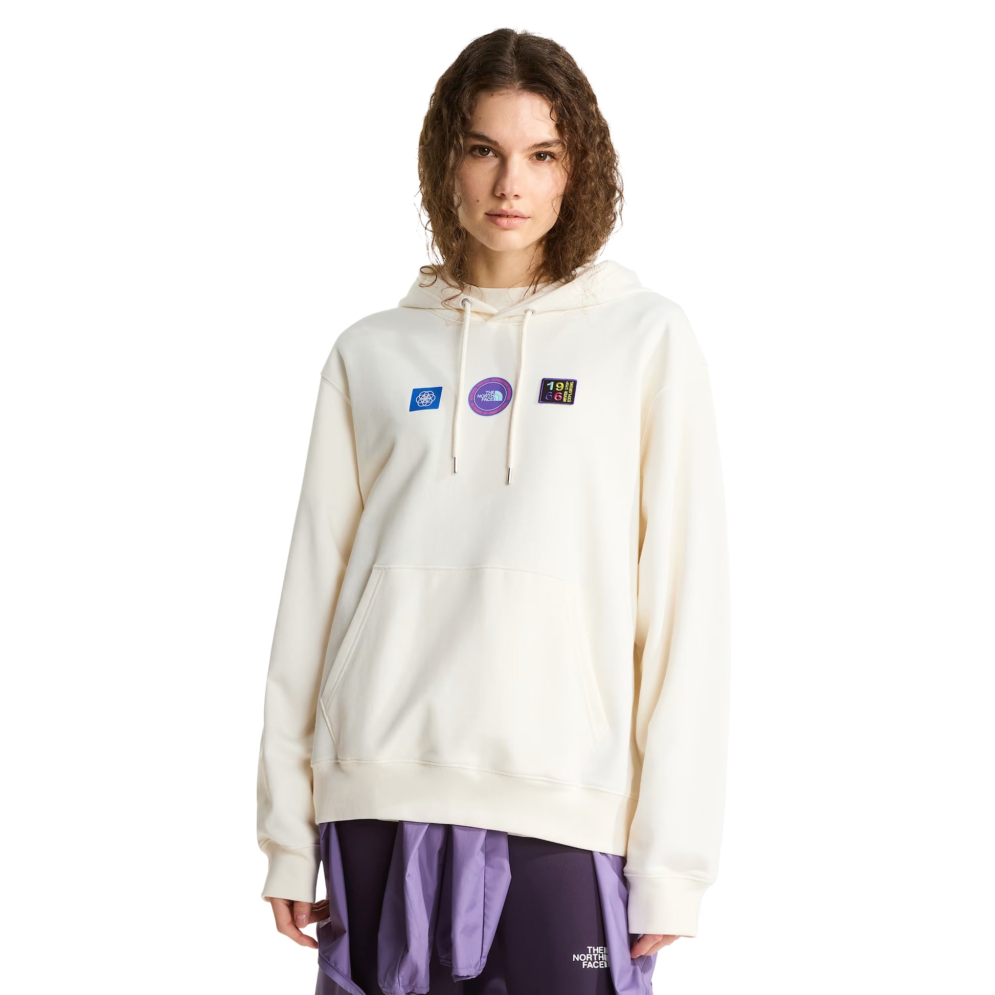 Hoodie unissexo The North Face Explorer Badges Relaxed em White Dune — modelo feminino de frente a 3/4, cabelo encaracolado castanho, capuz levantado com cordão branco, três emblemas Explorer estampados em serigrafia à base de água no peito (azul quadrado com globo, circular roxo "The North Face", retangular escuro "1966 Never Stop Exploring" multicolor), bolso canguru visível, French Terry White Dune 360 g/m², fundo branco.