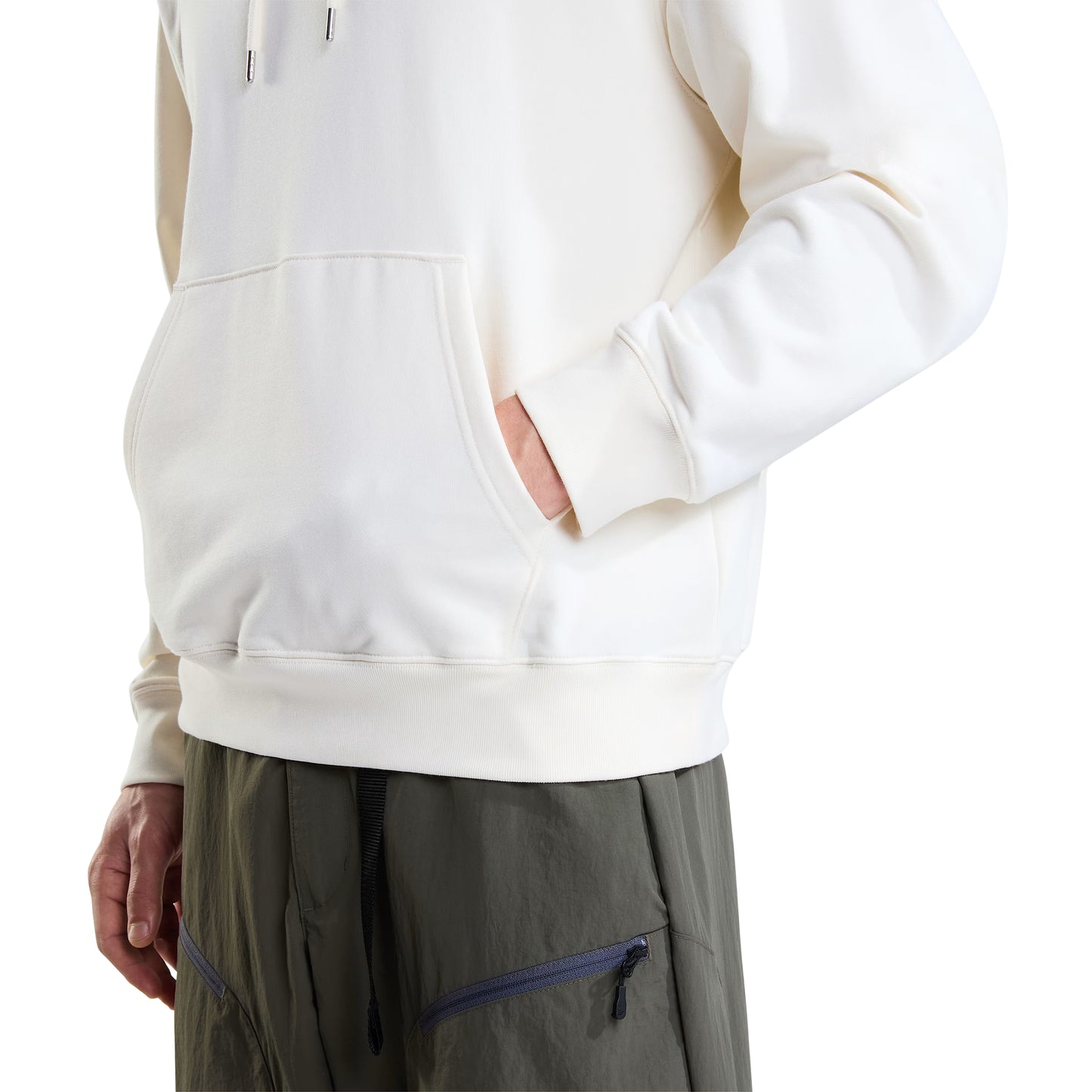 Hoodie unissexo The North Face Explorer Badges Relaxed em White Dune — modelo masculino, detalhe da parte inferior, meio corpo, calças cargo verde-oliva TNF, mão no bolso canguru, bainha canelada e punho canelado visíveis, French Terry White Dune 360 g/m², fundo branco.