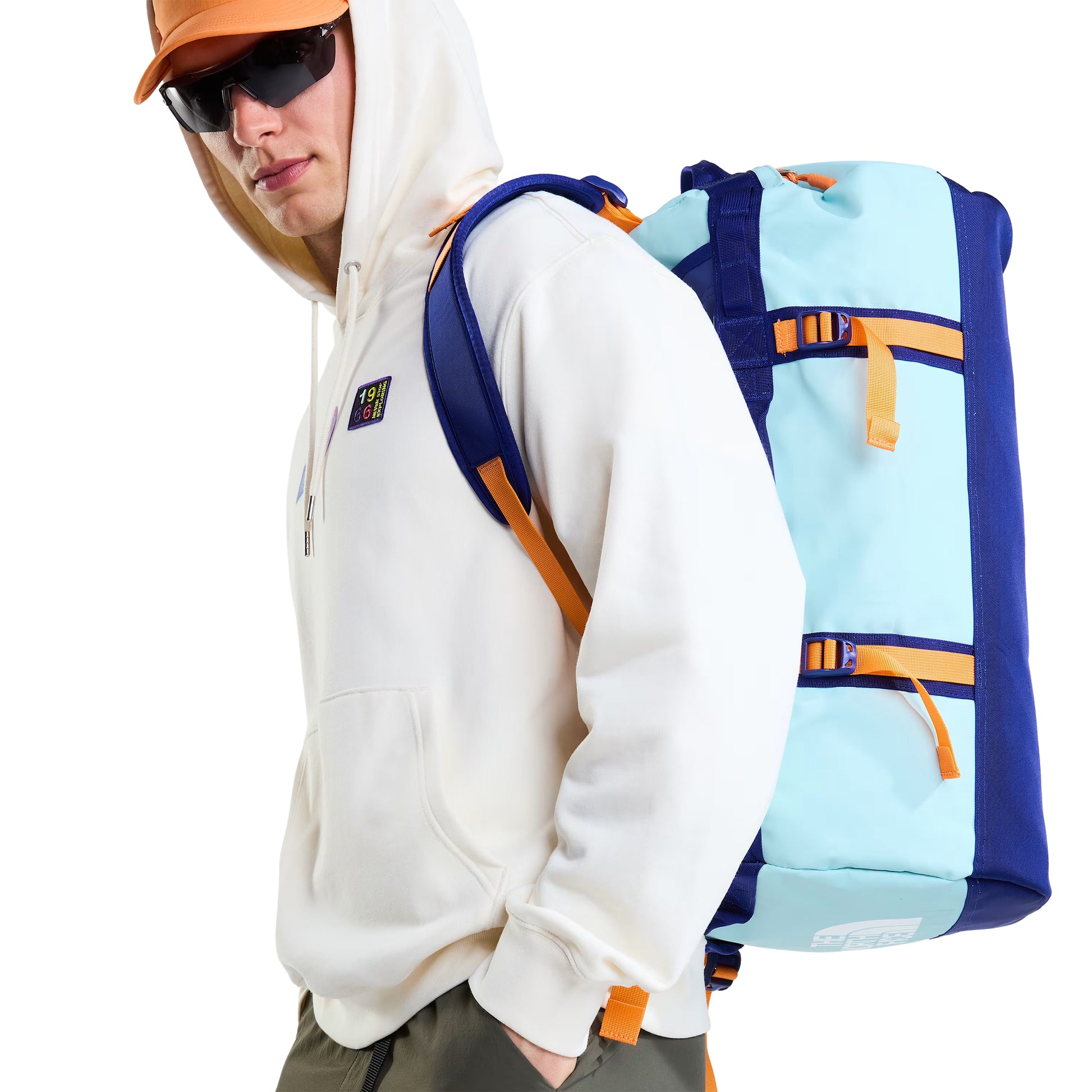 Hoodie unissexo The North Face Explorer Badges Relaxed em White Dune — modelo masculino de perfil esquerdo com mochila duffel TNF azul-claro e azul-marinho às costas, boné técnico laranja TNF, óculos de sol wrap escuros, calças cargo verde-oliva, capuz levantado, emblema "1966" parcialmente visível no peito, French Terry White Dune 360 g/m², fundo branco.