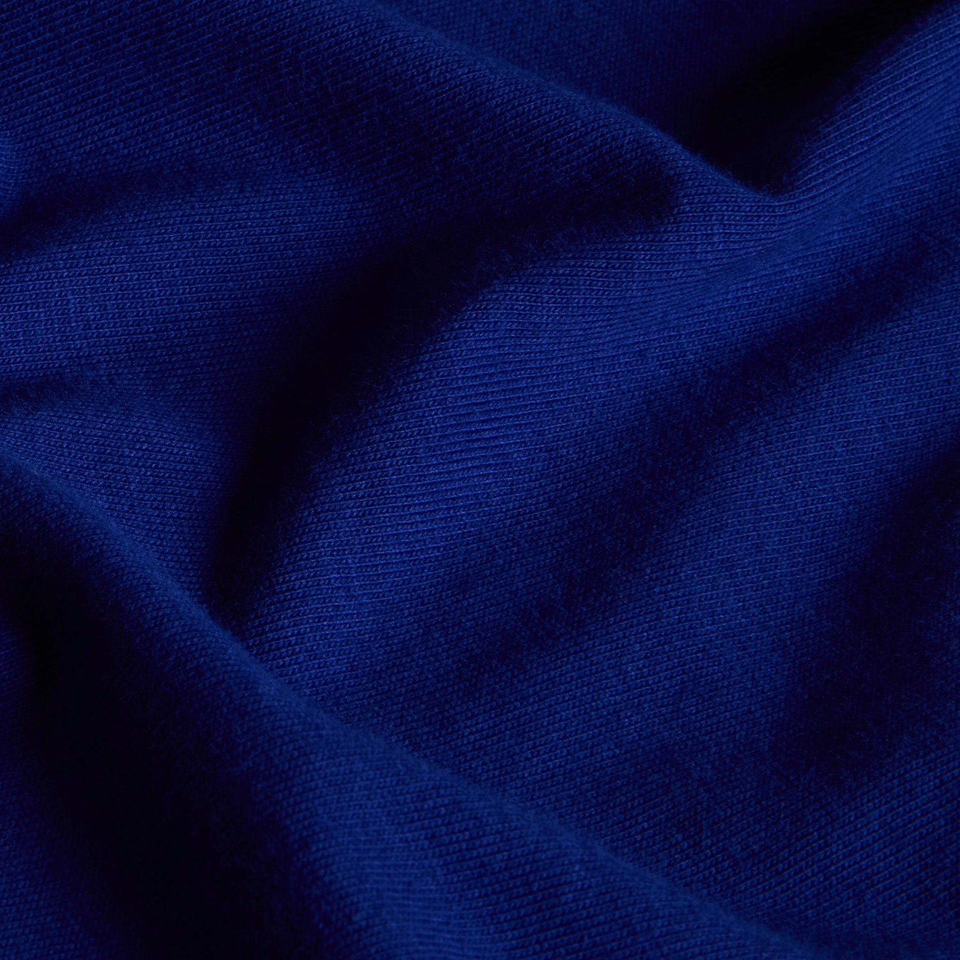 T-shirt unissexo The North Face Explorer Badges Relaxed em Bold Blue — detalhe da textura do jersey, close-up do tecido 100% algodão Bold Blue 220 g/m² com dobras naturais, textura macia e estrutura do jersey visíveis, fundo escuro.