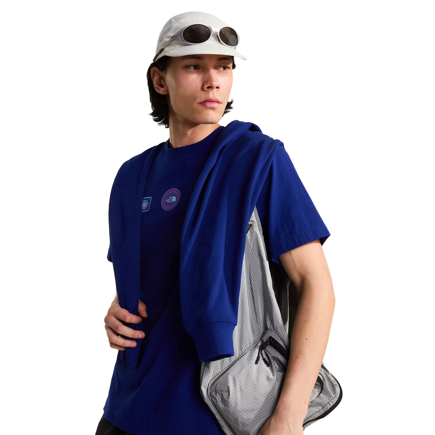 T-shirt unissexo The North Face Explorer Badges Relaxed em Bold Blue — modelo masculino de frente a 3/4, boné técnico branco com óculos de sol wrap brancos, mochila sling TNF cinzento claro ao ombro, casaco TNF Bold Blue ao braço, dois emblemas Explorer visíveis no peito (azul quadrado e circular roxo TNF), jersey Bold Blue e corte relaxado visíveis, fundo branco.