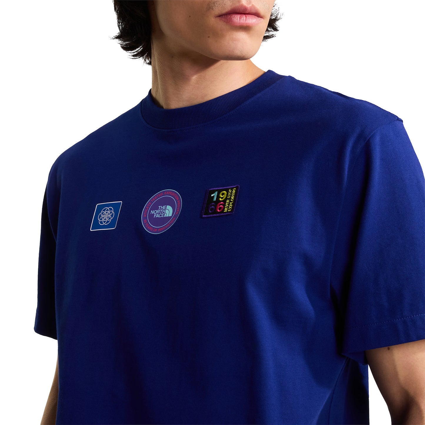 T-shirt unissexo The North Face Explorer Badges Relaxed em Bold Blue — modelo masculino de frente a 3/4, boné técnico branco com óculos de sol wrap brancos, mochila sling TNF cinzento claro ao ombro, casaco TNF Bold Blue ao braço, dois emblemas Explorer visíveis no peito (azul quadrado e circular roxo TNF), jersey Bold Blue e corte relaxado visíveis, fundo branco.
