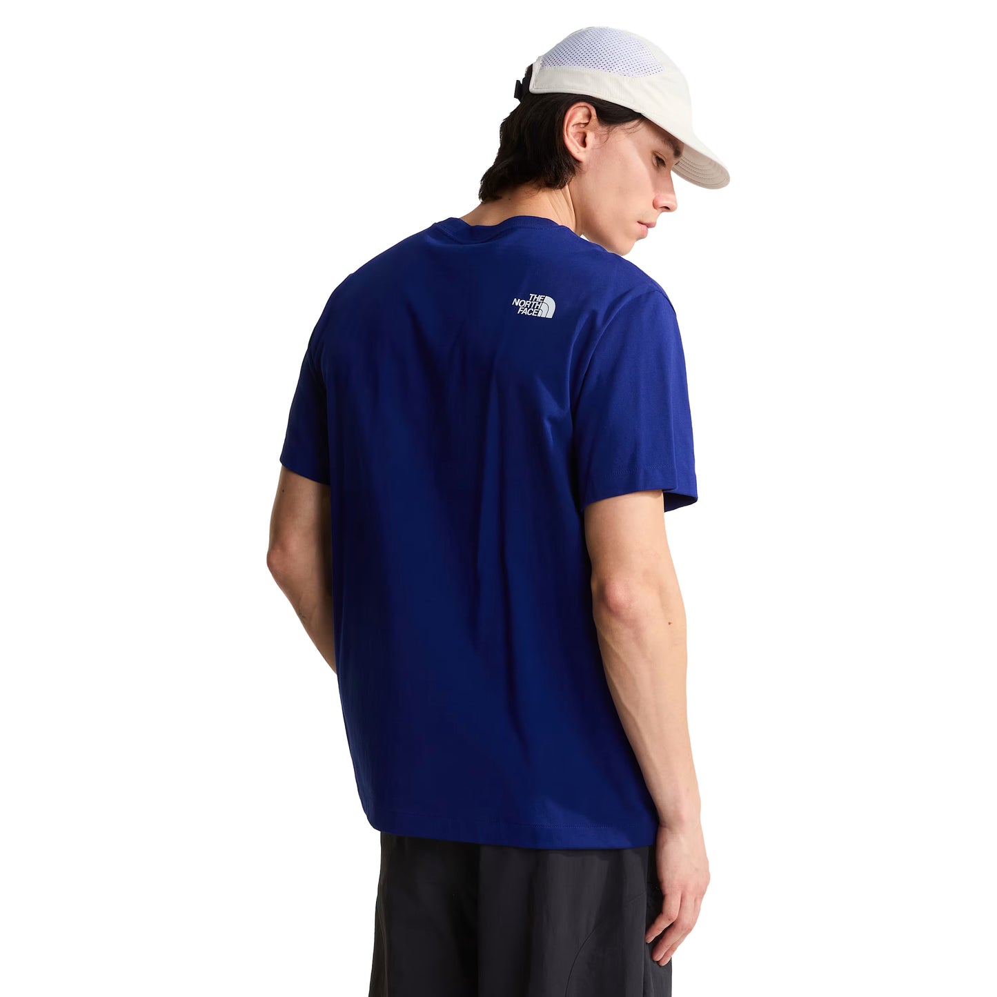 T-shirt unissexo The North Face Explorer Badges Relaxed em Bold Blue — modelo masculino de costas a 3/4, boné técnico branco de malha, cabelo escuro médio, calças cargo pretas largas, costas lisas em jersey Bold Blue, logótipo "The North Face" half-dome estampado em serigrafia à base de água no ombro direito nas costas, corte relaxado com ombros descaídos visíveis, fundo branco.
