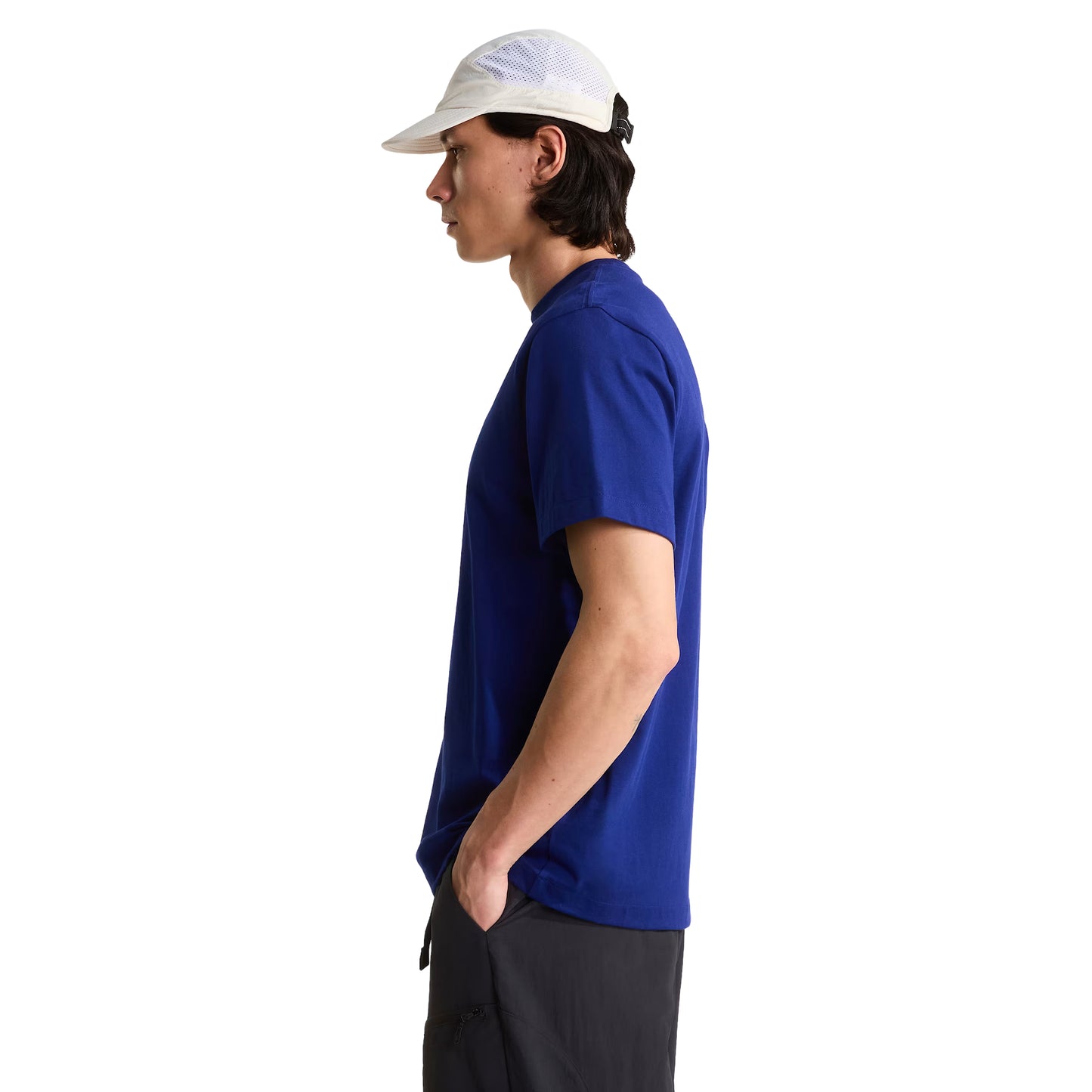 T-shirt unissexo The North Face Explorer Badges Relaxed em Bold Blue — modelo masculino de perfil esquerdo, boné técnico branco de malha, cabelo escuro médio, calças cargo pretas largas, corte relaxado com ombros descaídos e jersey Bold Blue 220 g/m² visíveis, emblemas Explorer parcialmente visíveis no peito, fundo branco.