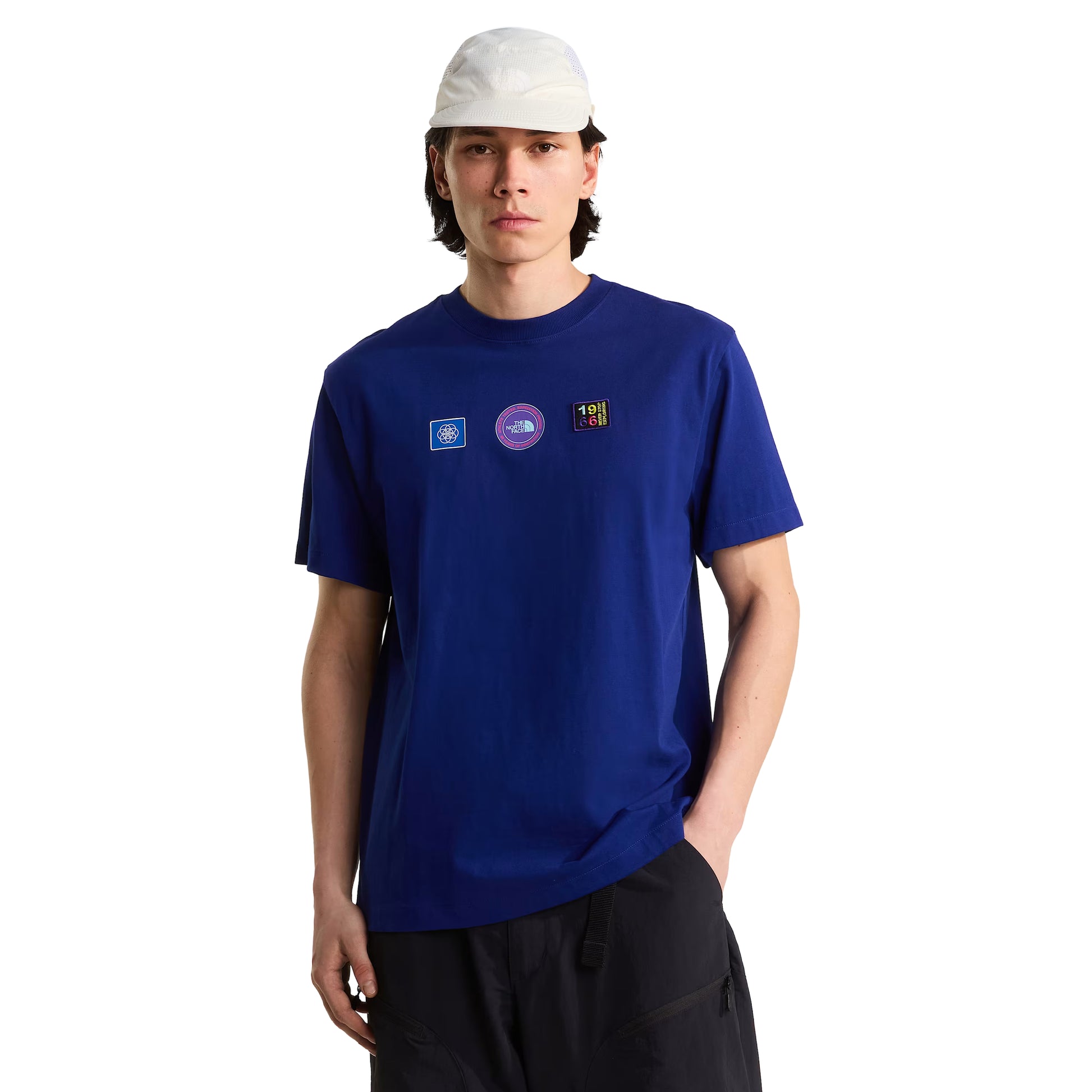 T-shirt unissexo The North Face Explorer Badges Relaxed em Bold Blue — modelo masculino de frente, meio corpo, boné técnico branco de malha, cabelo escuro médio, calças cargo pretas largas, três emblemas Explorer estampados em serigrafia à base de água no peito (azul quadrado com globo, circular roxo "The North Face", retangular escuro "1966 Never Stop Exploring" multicolor), gola canelada e jersey Bold Blue 220 g/m² visíveis, fundo branco.