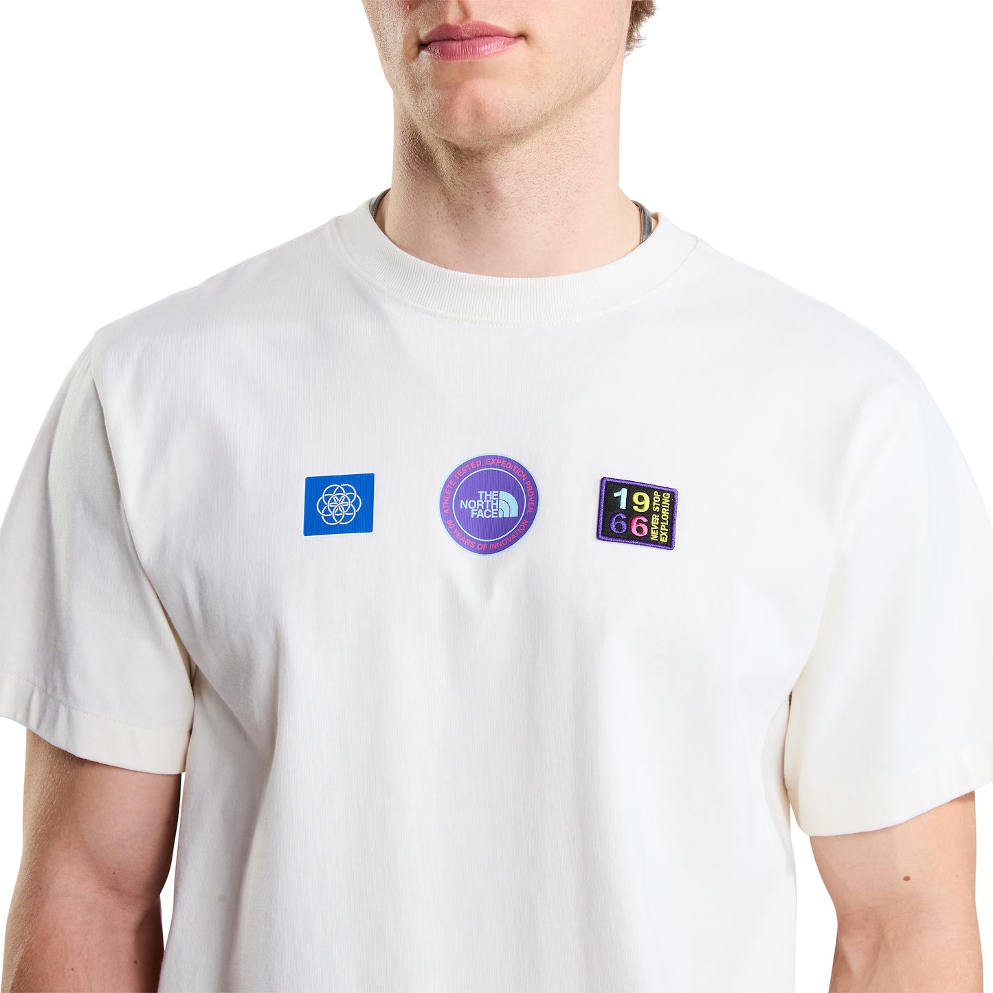 T-shirt unissexo The North Face Explorer Badges Relaxed em White Dune — modelo masculino de frente, meio corpo, detalhe do peito, três emblemas Explorer estampados em serigrafia à base de água em close-up: emblema azul quadrado com globo à esquerda, emblema circular roxo "The North Face" ao centro, emblema retangular escuro "1966 Never Stop Exploring" com letras multicolor (vermelho, amarelo, verde, azul) à direita, gola canelada e jersey White Dune 220 g/m² visíveis, fundo branco.