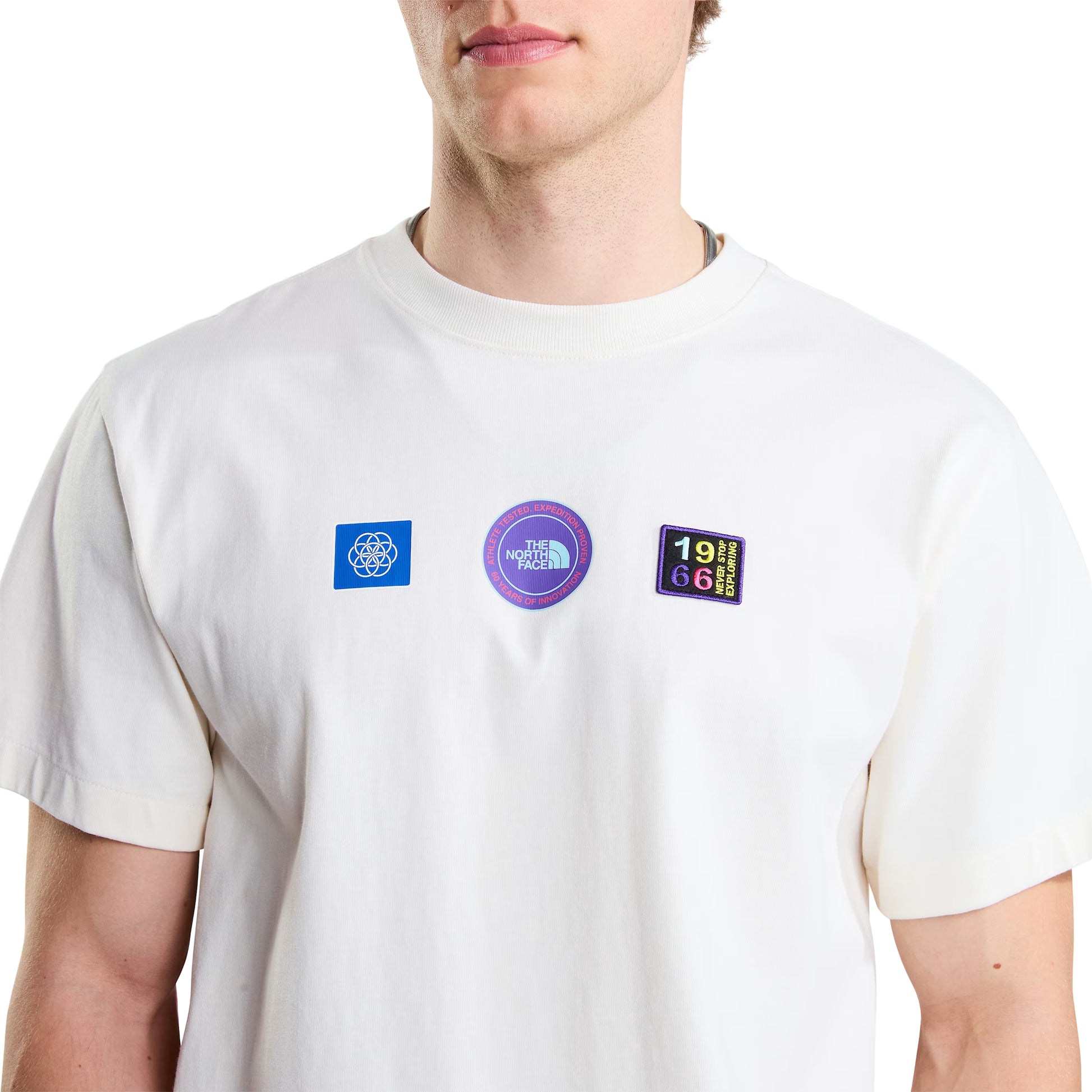 T-shirt unissexo The North Face Explorer Badges Relaxed em White Dune — modelo masculino de frente, meio corpo, detalhe do peito, três emblemas Explorer estampados em serigrafia à base de água em close-up: emblema azul quadrado com globo à esquerda, emblema circular roxo "The North Face" ao centro, emblema retangular escuro "1966 Never Stop Exploring" com letras multicolor (vermelho, amarelo, verde, azul) à direita, gola canelada e jersey White Dune 220 g/m² visíveis, fundo branco.