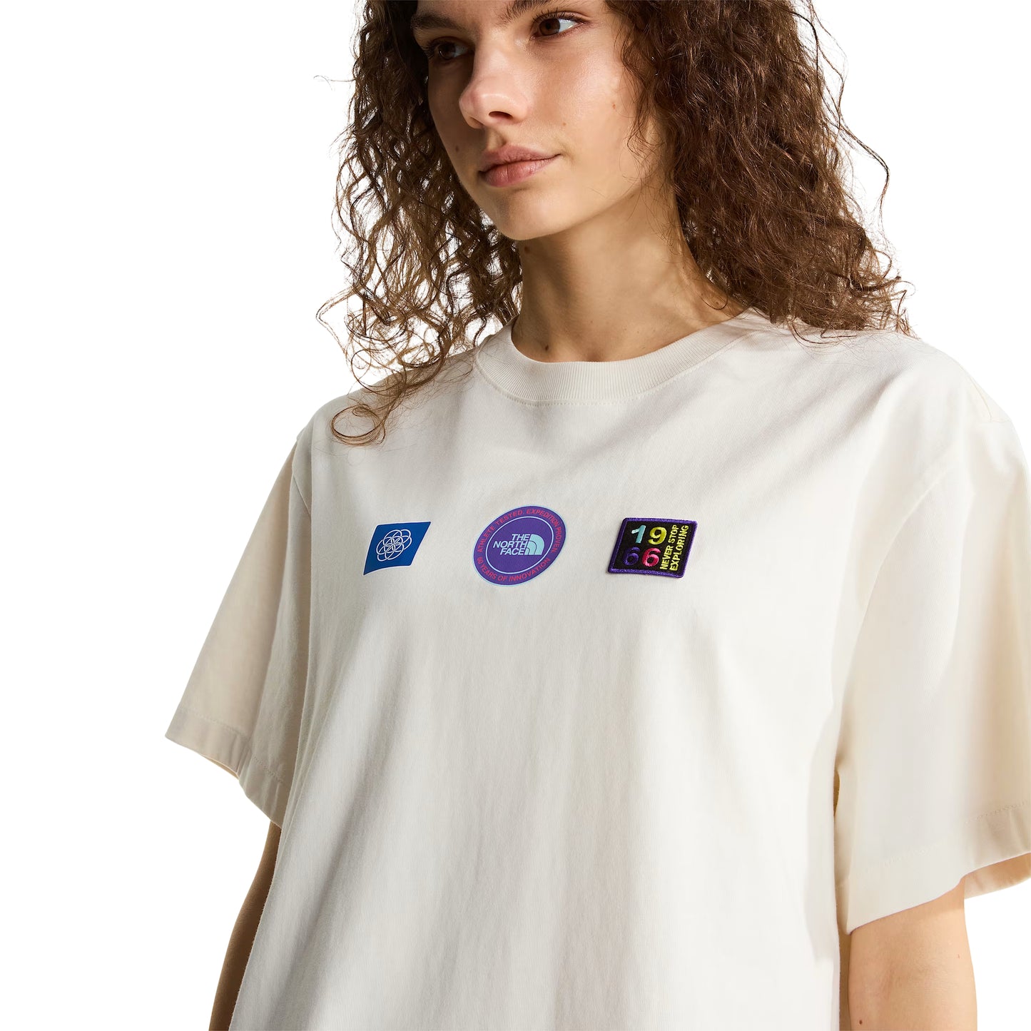 T-shirt unissexo The North Face Explorer Badges Relaxed em White Dune — modelo feminino de frente, meio corpo, cabelo encaracolado solto, detalhe do peito, três emblemas Explorer estampados em serigrafia à base de água em close-up: emblema azul quadrado com globo à esquerda, emblema circular roxo "The North Face" ao centro, emblema retangular escuro "1966 Never Stop Exploring" com letras multicolor à direita, gola canelada e jersey White Dune 220 g/m² visíveis, fundo branco.