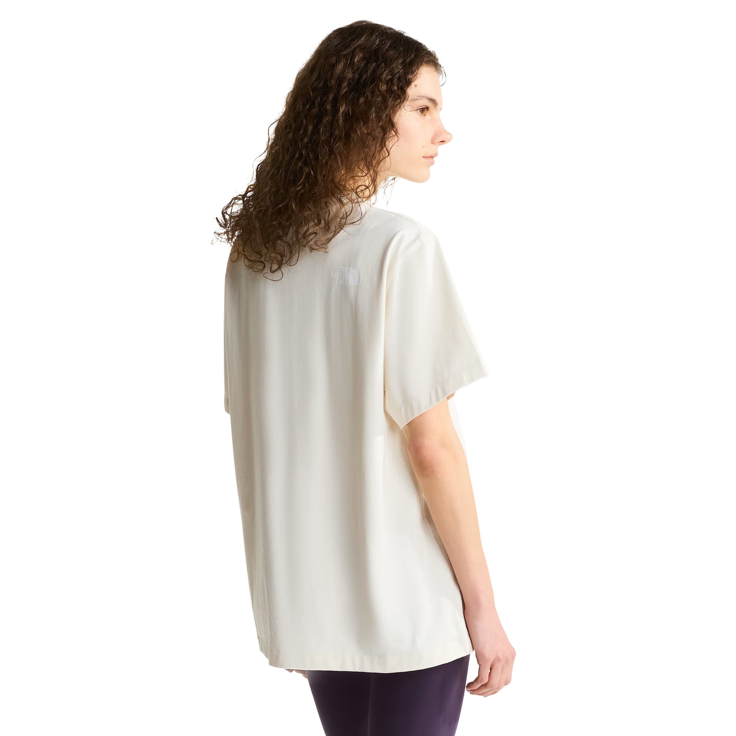 T-shirt unissexo The North Face Explorer Badges Relaxed em White Dune — modelo feminino de costas a 3/4, cabelo encaracolado solto, leggings roxo escuro, costas lisas em jersey White Dune, logótipo TNF estampado em serigrafia à base de água no ombro direito nas costas, corte relaxado com comprimento alongado visível, fundo branco.