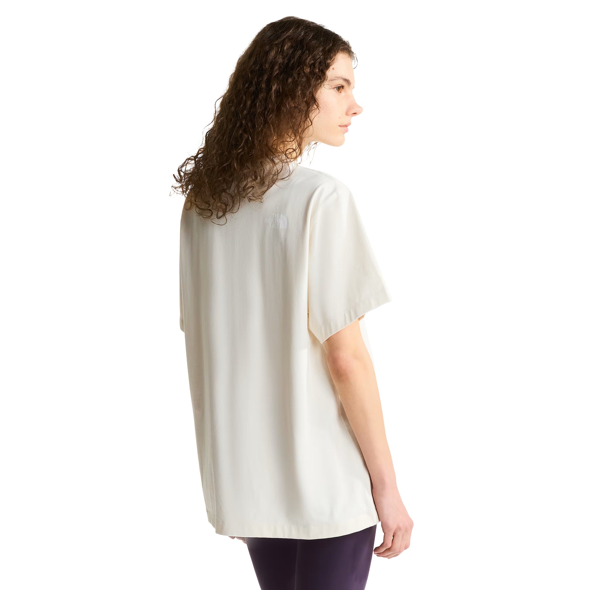 T-shirt unissexo The North Face Explorer Badges Relaxed em White Dune — modelo feminino de costas a 3/4, cabelo encaracolado solto, leggings roxo escuro, costas lisas em jersey White Dune, logótipo TNF estampado em serigrafia à base de água no ombro direito nas costas, corte relaxado com comprimento alongado visível, fundo branco.
