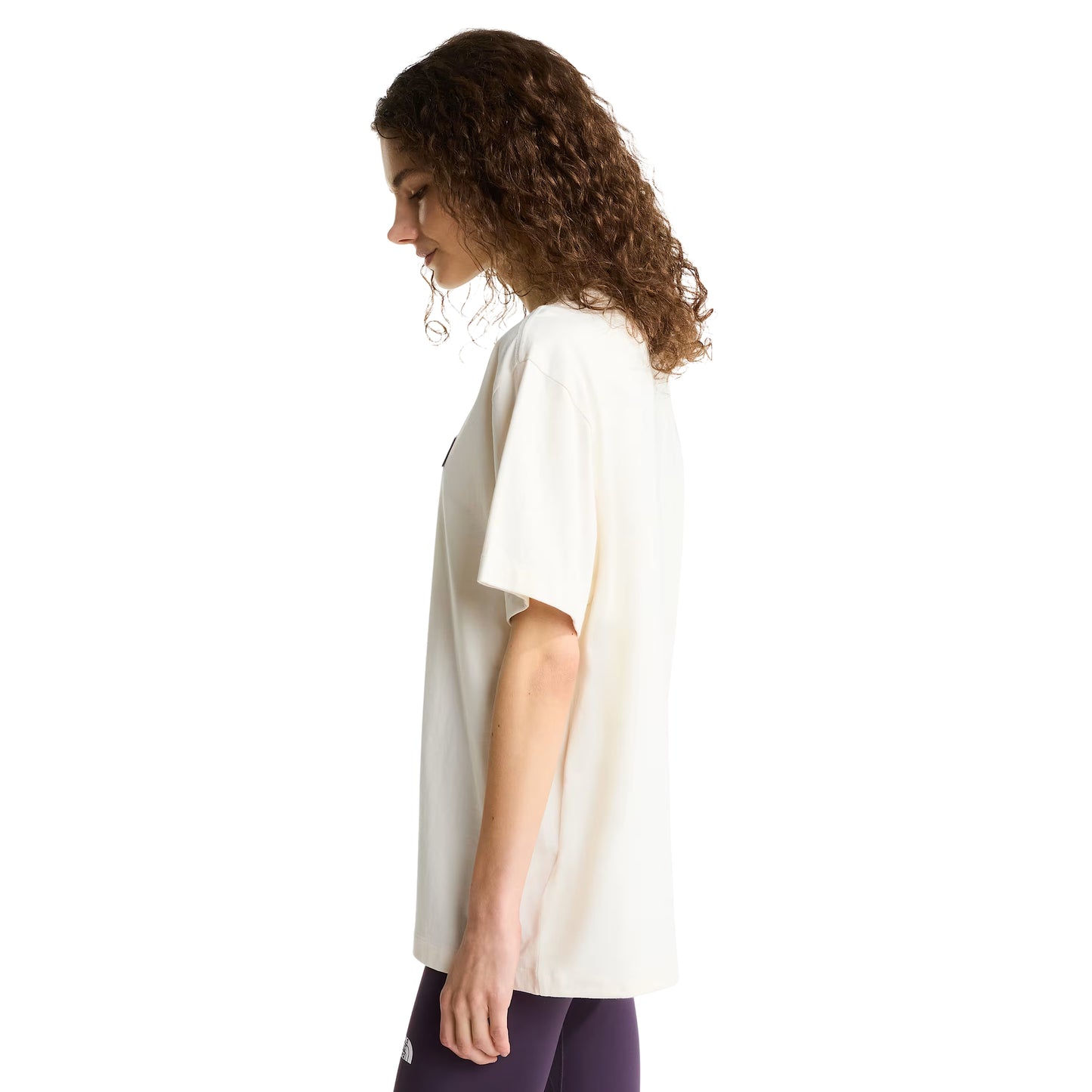 T-shirt unissexo The North Face Explorer Badges Relaxed em White Dune — modelo feminino de perfil direito, cabelo encaracolado solto, leggings roxo escuro, emblemas Explorer parcialmente visíveis no peito, corte relaxado com ombros descaídos e comprimento alongado visíveis, fundo branco.