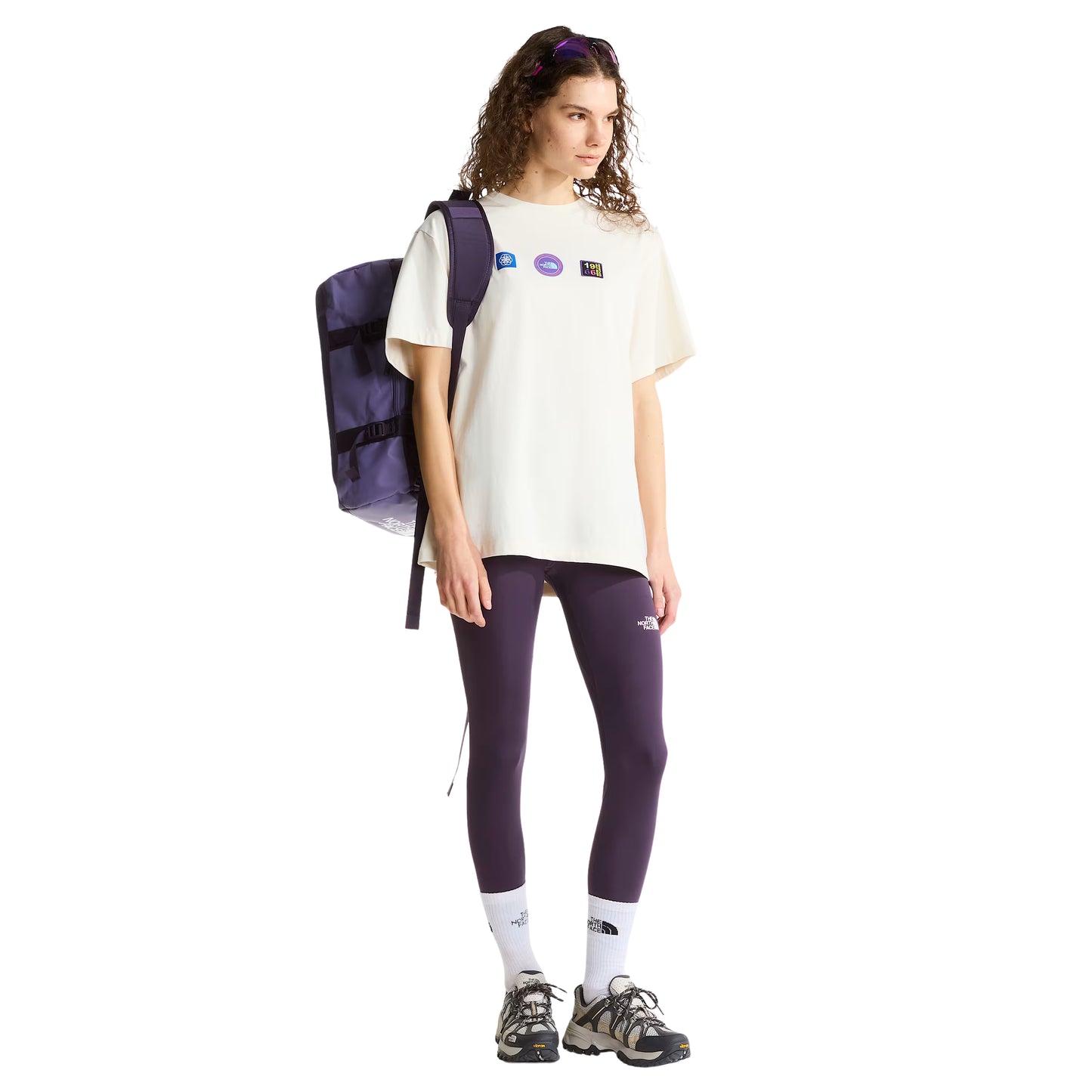 T-shirt unissexo The North Face Explorer Badges Relaxed em White Dune — modelo feminino de frente, corpo inteiro, óculos de sol roxos na cabeça, mochila TNF roxa às costas, leggings roxo escuro TNF, meias brancas TNF, sapatilhas de trail, três emblemas Explorer estampados no peito visíveis, corte relaxado com comprimento alongado visível, fundo branco.