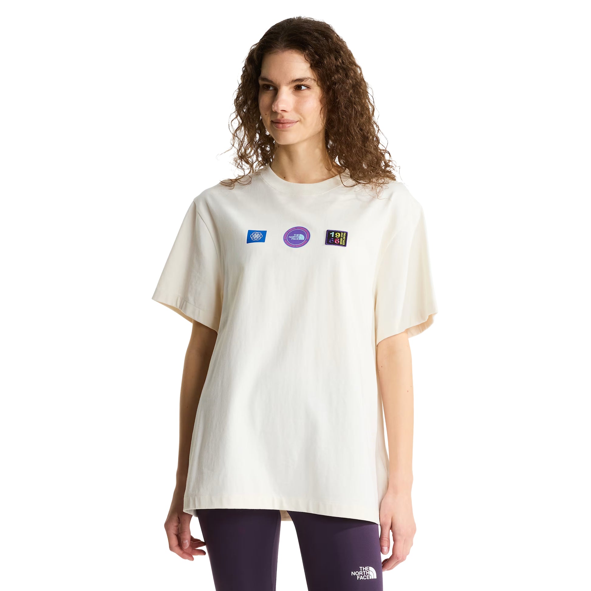 T-shirt unissexo The North Face Explorer Badges Relaxed em White Dune — modelo feminino de frente, meio corpo, cabelo encaracolado solto, leggings roxo escuro, três emblemas Explorer estampados em serigrafia à base de água no peito (azul retangular, roxo circular TNF, multicolor "1966"), gola canelada e jersey White Dune 220 g/m² visíveis, fundo branco.