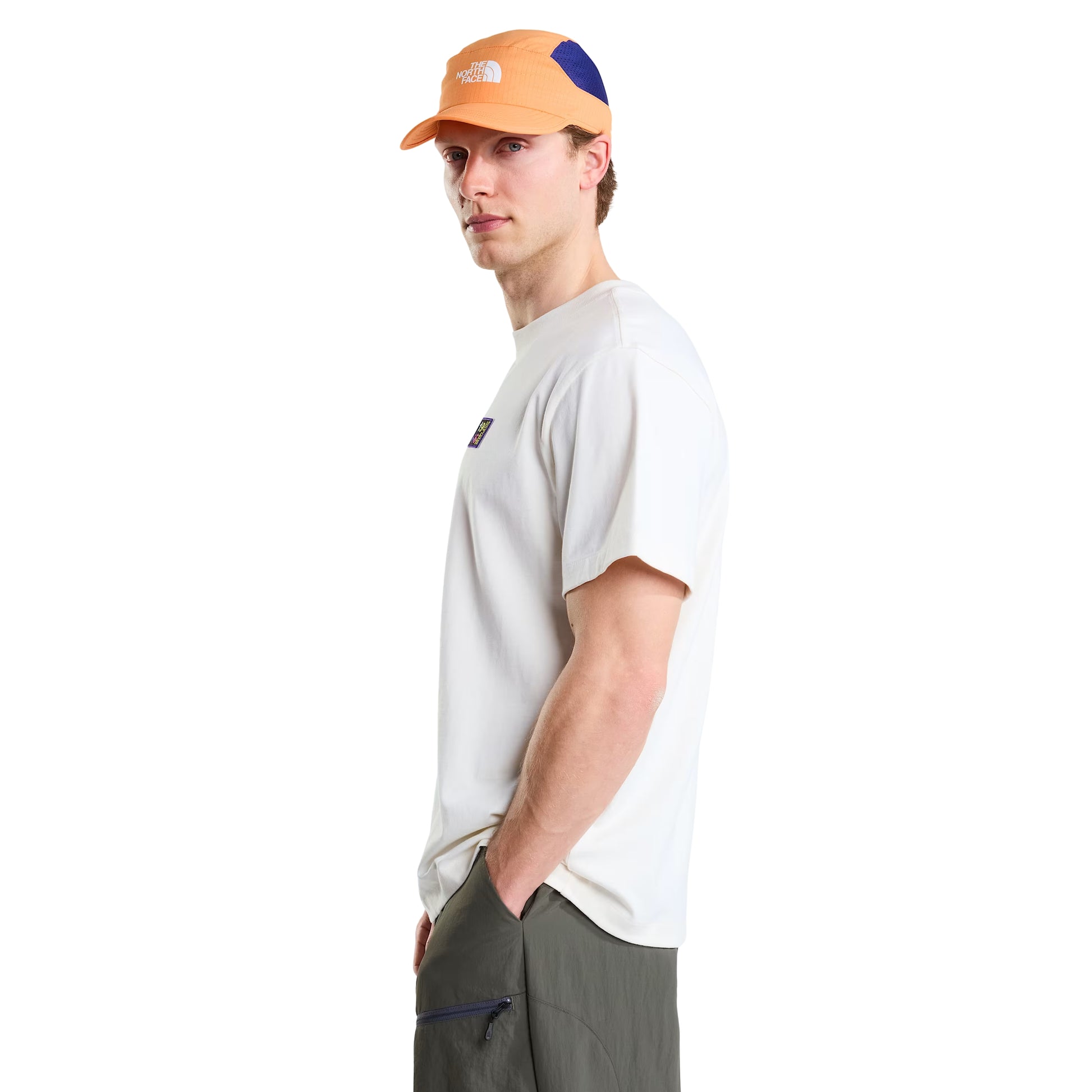T-shirt unissexo The North Face Explorer Badges Relaxed em White Dune — modelo masculino de perfil direito, boné técnico laranja e azul TNF, calções cargo verde-oliva, logótipo TNF estampado em serigrafia à base de água no ombro direito visível, corte relaxado com ombros descaídos e jersey White Dune 220 g/m² visíveis, fundo branco.
