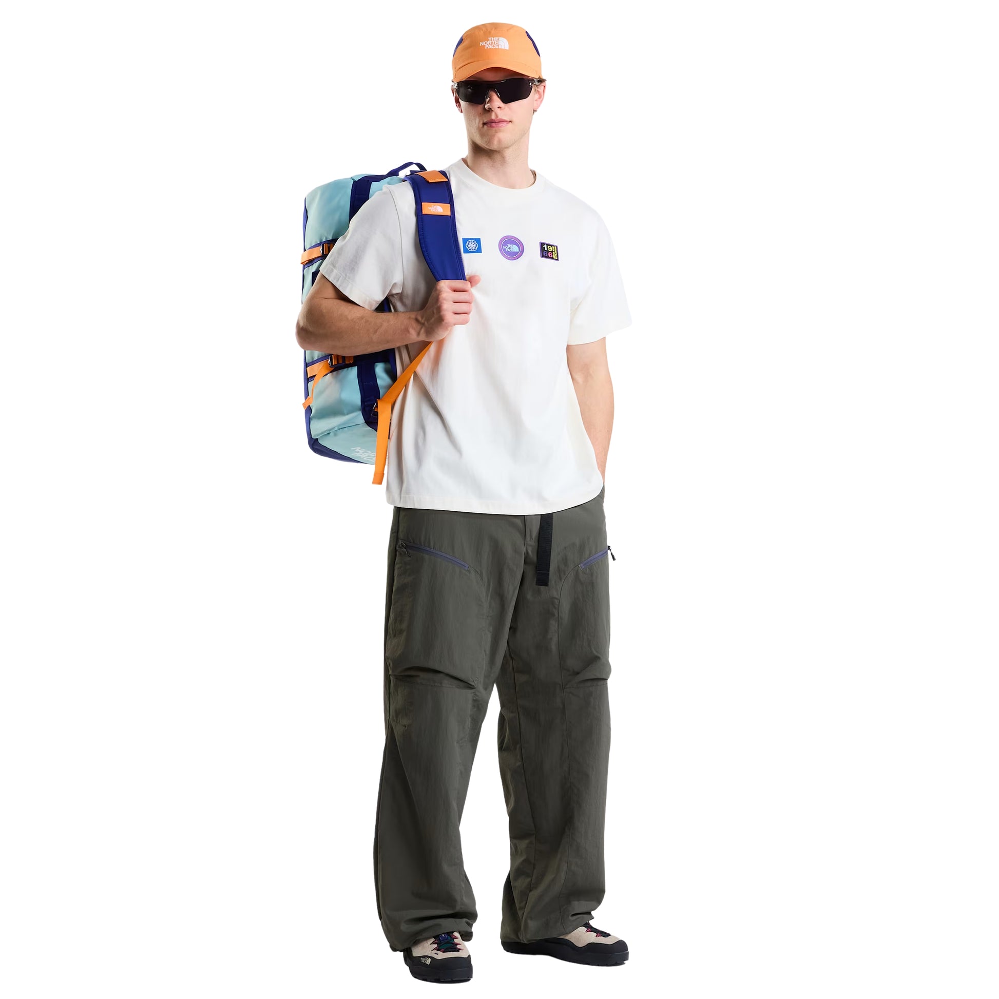 T-shirt unissexo The North Face Explorer Badges Relaxed em White Dune — modelo masculino de frente, corpo inteiro, boné técnico laranja TNF, óculos de sol escuros, mochila duffel azul e azul-claro TNF ao ombro, calças cargo verde-oliva largas, sapatilhas de trail, três emblemas Explorer estampados no peito visíveis, corte relaxado com ombros descaídos e jersey White Dune visíveis, fundo branco.