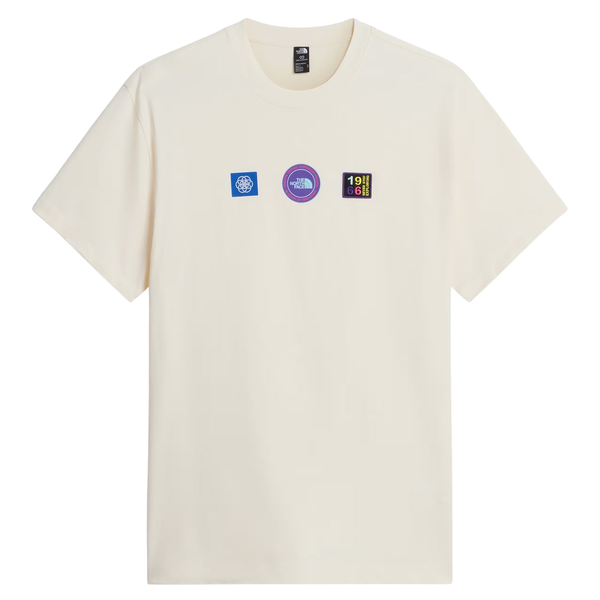 T-shirt unissexo The North Face Explorer Badges Relaxed em White Dune — vista frontal, produto sem modelo, três emblemas Explorer estampados em serigrafia à base de água no peito: emblema azul quadrado com globo à esquerda, emblema circular roxo "The North Face" ao centro, emblema retangular escuro "1966 Never Stop Exploring" com letras multicolor à direita, etiqueta TNF na gola interior visível, jersey White Dune 220 g/m² e gola canelada visíveis, fundo branco.