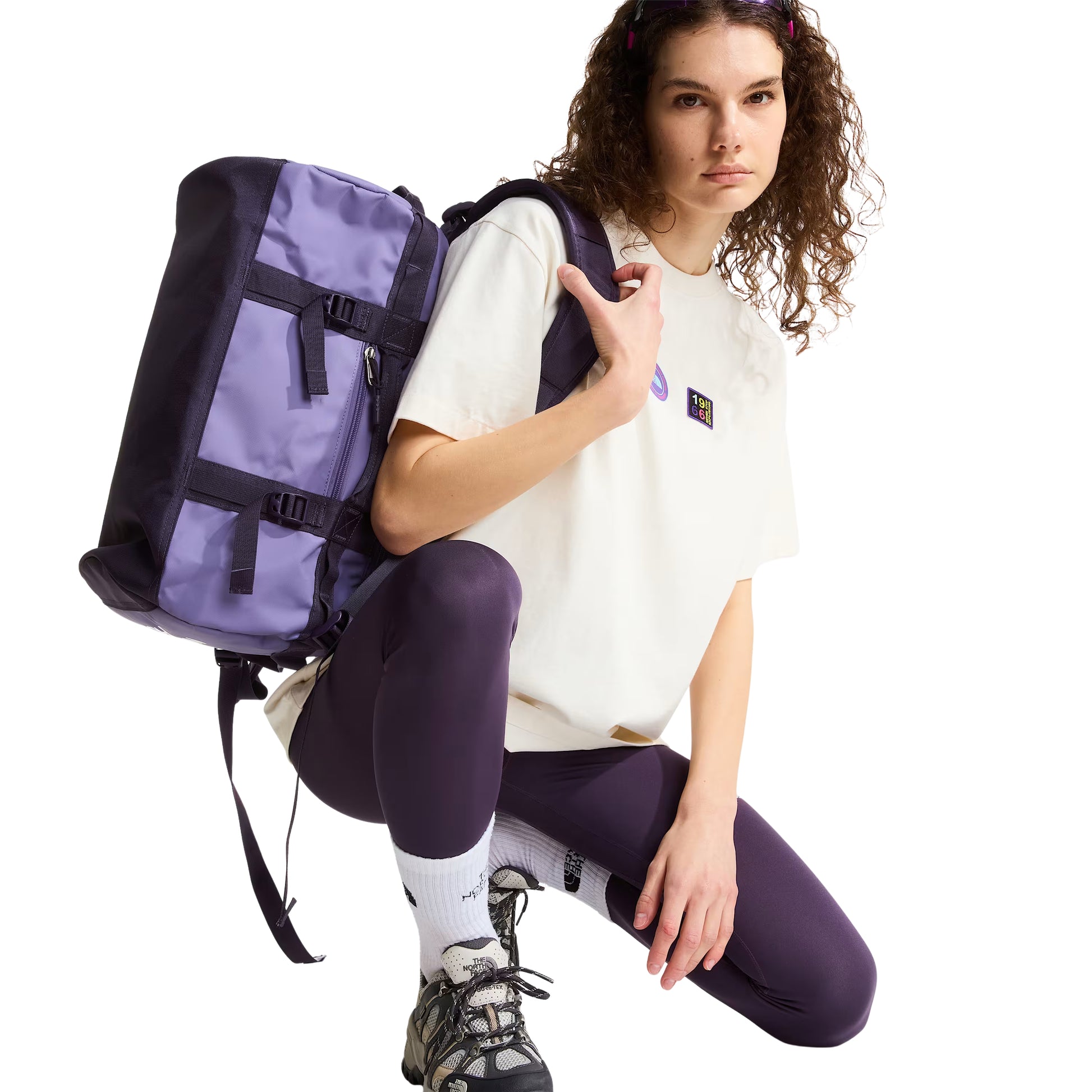 T-shirt unissexo The North Face Explorer Badges Relaxed em White Dune — modelo feminino de frente, meio corpo, cabelo encaracolado solto, mochila TNF roxa e preta às costas, leggings roxo escuro, botas de hiking TNF GORE-TEX, meias brancas TNF, dois emblemas Explorer visíveis no peito (circular roxo "The North Face" e retangular escuro "1966" multicolor), corte relaxado com ombros descaídos e jersey White Dune visíveis, fundo branco.
