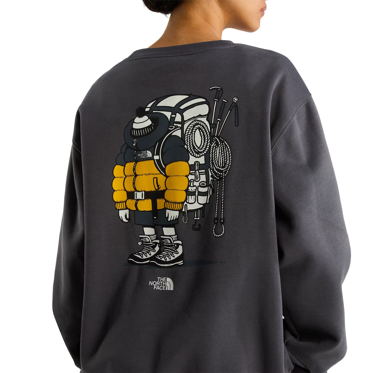 Sweatshirt unissexo The North Face Gear Up Get Out Relaxed Crew em Anthracite Grey — modelo feminino de costas, meio corpo superior, polar cinzento antracite, detalhe do gráfico Gear Up Get Out em serigrafia à base de água nas costas com personagem de alpinista totalmente equipado em amarelo e cinzento — casaco puffer amarelo, mochila, boné de malha, corda de escalada, picaretas e botas TNF —, logo The North Face em branco por baixo, fundo branco.