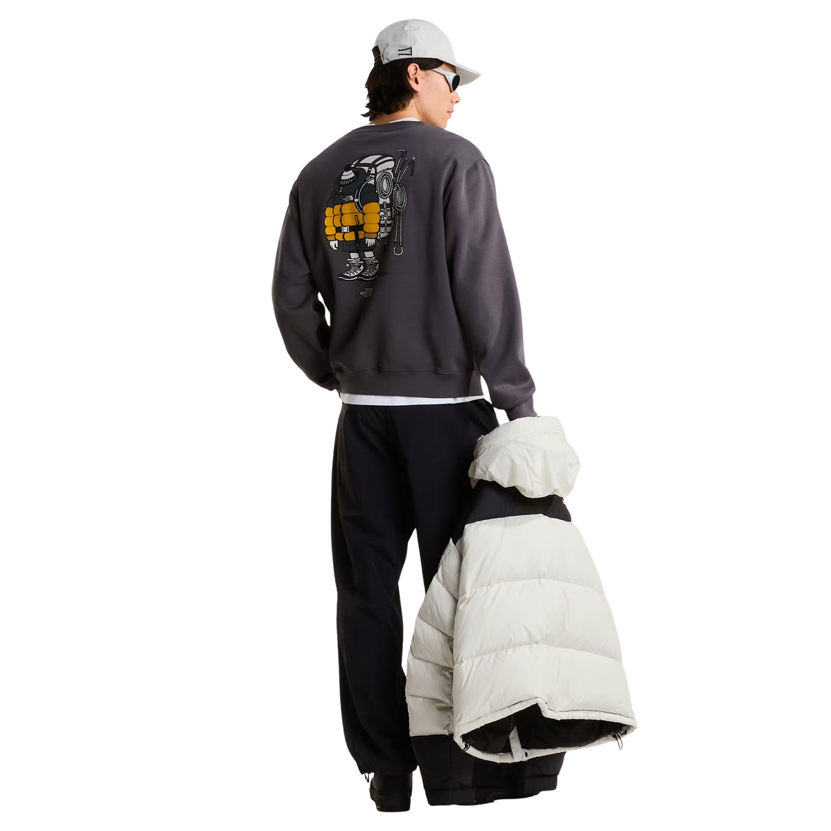 Sweatshirt unissexo The North Face Gear Up Get Out Relaxed Crew em Anthracite Grey — modelo masculino de costas, corpo inteiro, polar cinzento antracite, gráfico Gear Up Get Out em serigrafia à base de água com personagem de equipamento TNF em amarelo nas costas, boné cinzento claro, óculos de sol escuros, casaco puffer branco seguro na mão direita, calças pretas largas, fundo branco.