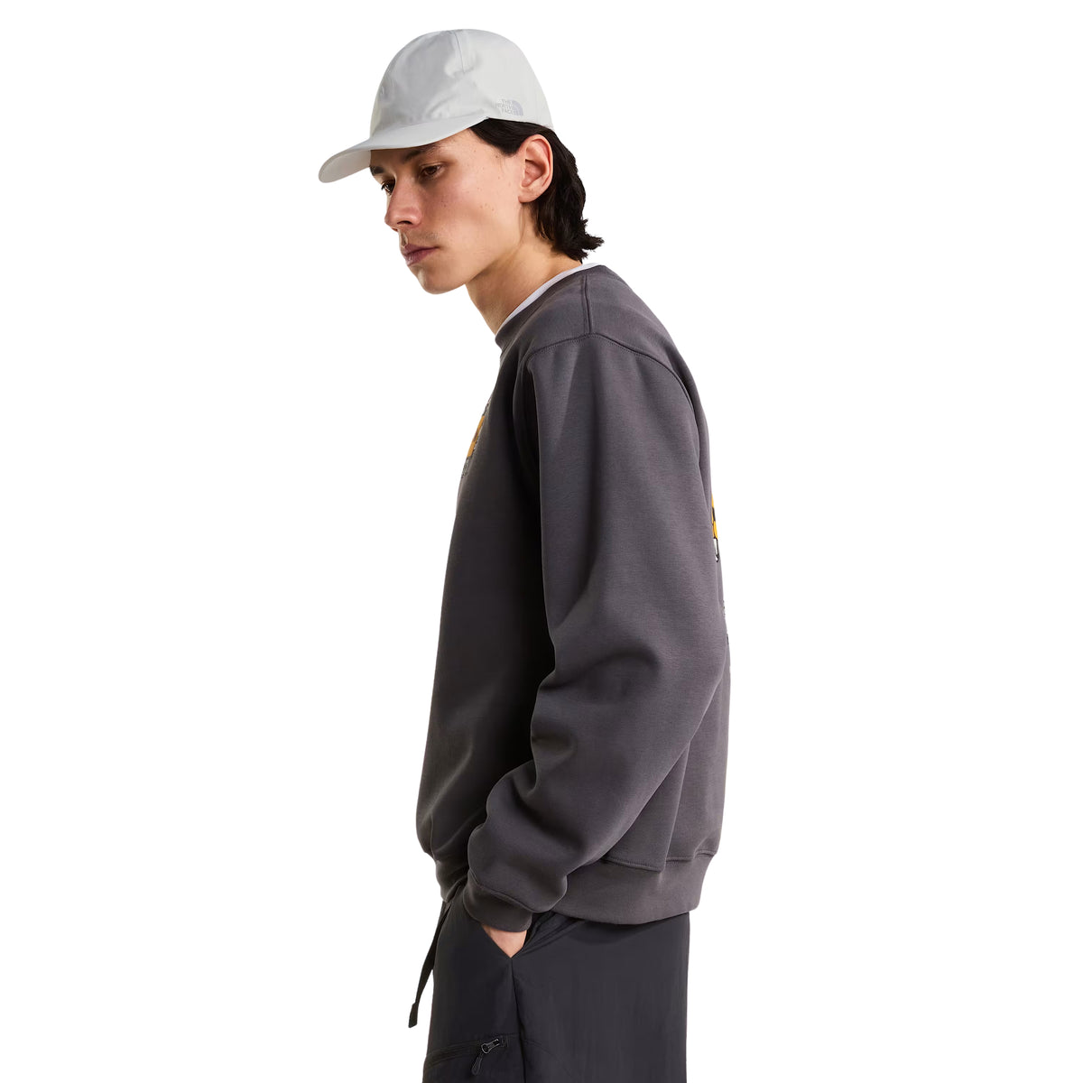 Sweatshirt unissexo The North Face Gear Up Get Out Relaxed Crew em Anthracite Grey — modelo masculino de perfil direito, corpo inteiro, polar cinzento antracite, gráfico Gear Up Get Out em serigrafia à base de água parcialmente visível no peito esquerdo, boné cinzento claro, mão no bolso, corte relaxed com ombros descaídos, calças cargo antracite, fundo branco.