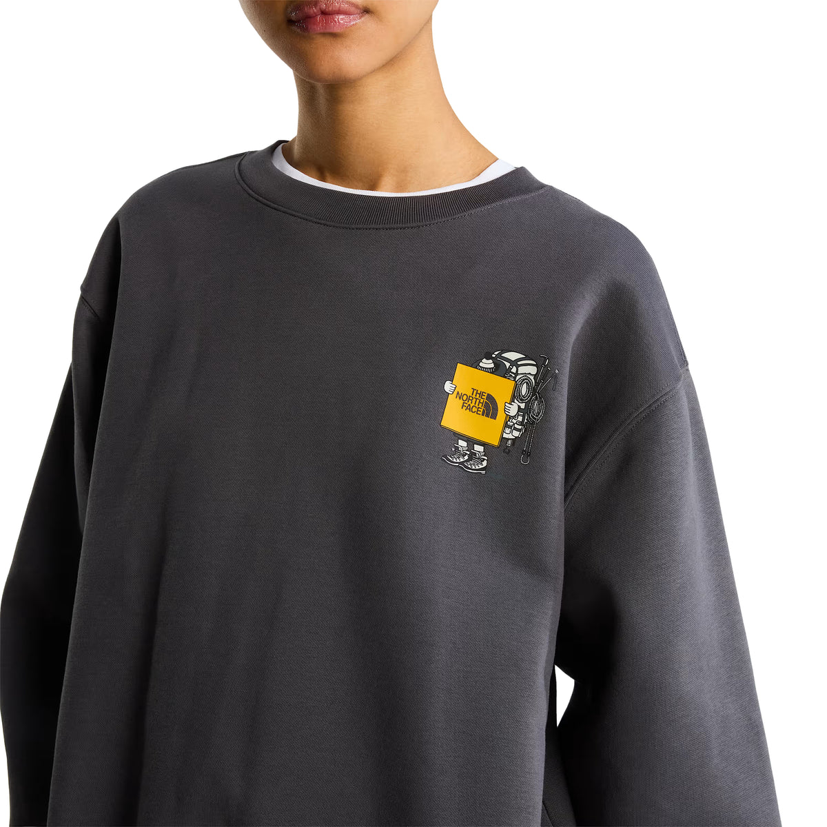 Sweatshirt unissexo The North Face Gear Up Get Out Relaxed Crew em Anthracite Grey — pormenor de meio corpo superior, modelo feminino de frente, polar cinzento antracite, gráfico Gear Up Get Out em serigrafia à base de água no peito esquerdo com personagem de equipamento TNF segurando placa amarela "The North Face", gola redonda canelada com t-shirt branca por baixo, fundo branco.
