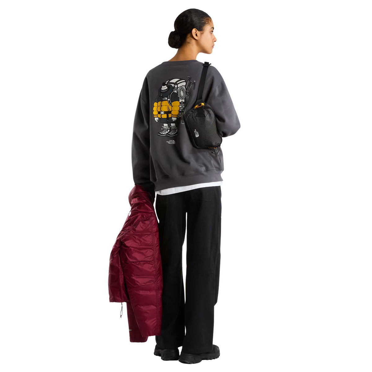 Sweatshirt unissexo The North Face Gear Up Get Out Relaxed Crew em Anthracite Grey — modelo feminino de costas, corpo inteiro, polar cinzento antracite, gráfico Gear Up Get Out em serigrafia à base de água com personagem de equipamento TNF em amarelo nas costas, saco crossbody preto TNF pendurado no ombro, casaco vermelho puffer seguro na mão esquerda, calças pretas largas, botas pretas, fundo branco.