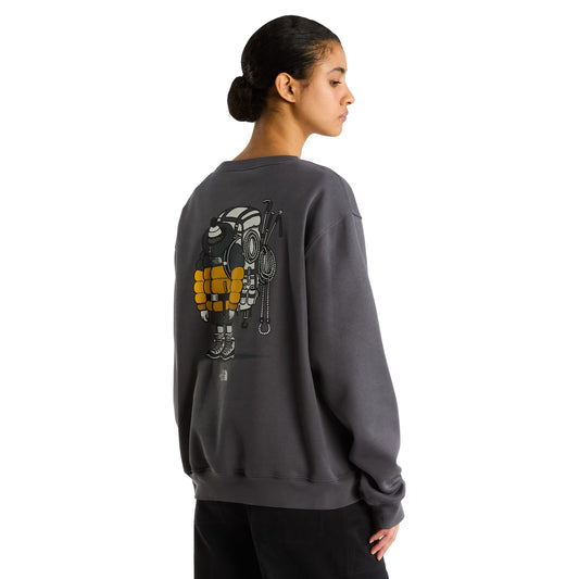 Sweatshirt unissexo The North Face Gear Up Get Out Relaxed Crew em Anthracite Grey — modelo feminino de costas, perfil esquerdo, corpo inteiro, polar cinzento antracite, gráfico Gear Up Get Out em serigrafia à base de água com equipamento de montanha colorido nas costas, cabelo apanhado, corte relaxed com ombros descaídos, calças pretas, fundo branco.