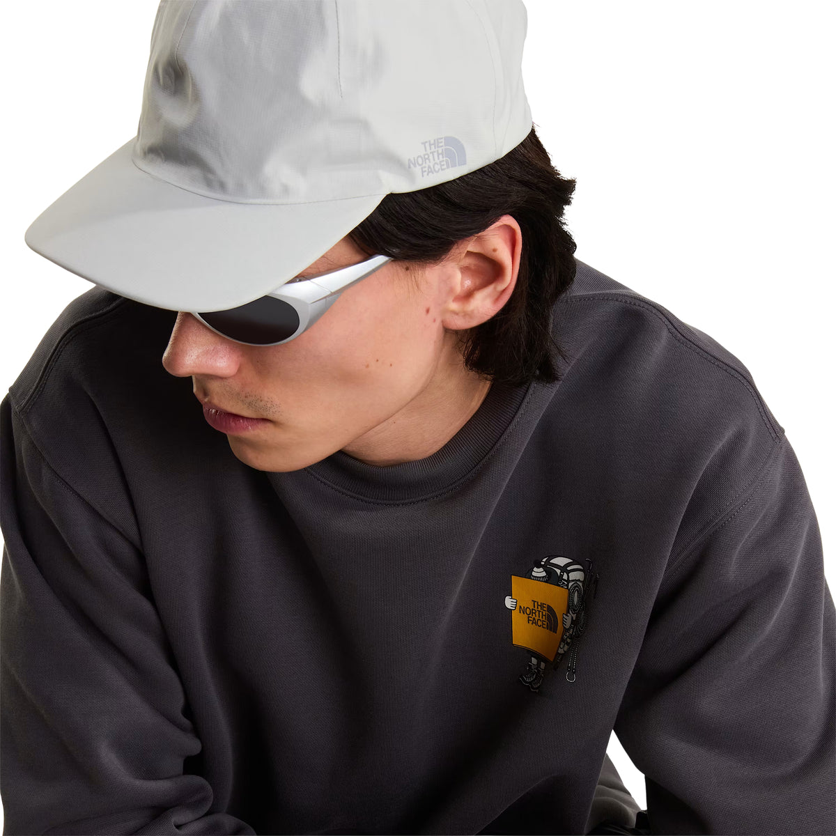 Sweatshirt unissexo The North Face Gear Up Get Out Relaxed Crew em Anthracite Grey — pormenor de meio corpo superior, modelo masculino inclinado para a frente, polar cinzento antracite, gráfico Gear Up Get Out em serigrafia à base de água no peito esquerdo com personagem de equipamento TNF, boné cinzento claro The North Face, óculos de sol wrap brancos, fundo branco.