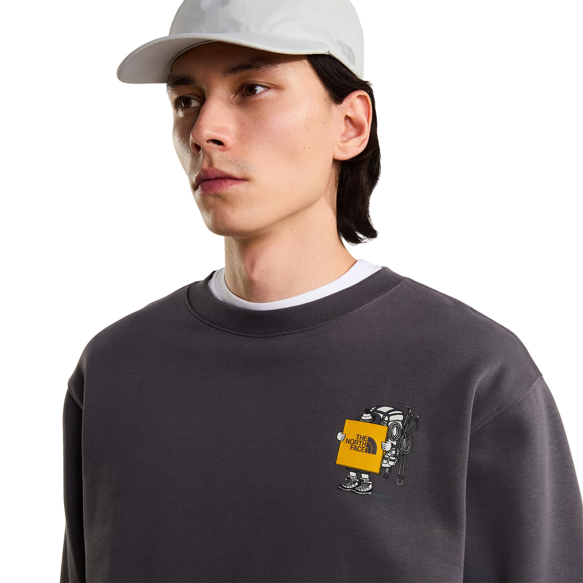 Sweatshirt unissexo The North Face Gear Up Get Out Relaxed Crew em Anthracite Grey — pormenor de meio corpo superior, modelo masculino de três quartos de frente, polar cinzento antracite, gráfico Gear Up Get Out em serigrafia à base de água no peito esquerdo com personagem de equipamento TNF segurando placa amarela "The North Face", boné cinzento claro The North Face, gola redonda canelada com t-shirt branca por baixo, fundo branco.