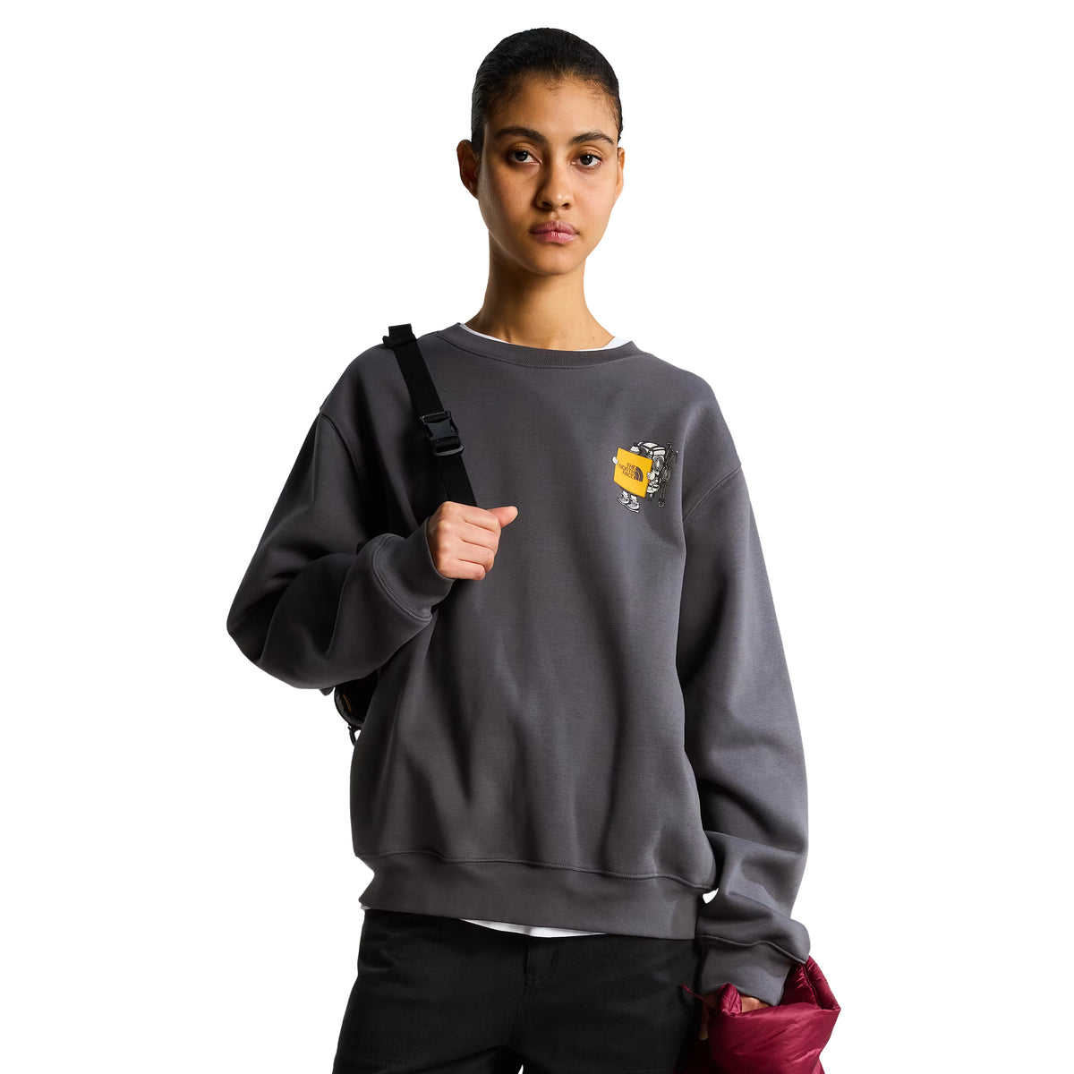 Sweatshirt unissexo The North Face Gear Up Get Out Relaxed Crew em Anthracite Grey — modelo feminino de frente, meio corpo, polar cinzento antracite, gráfico Gear Up Get Out em serigrafia à base de água com personagem de equipamento TNF em amarelo no peito esquerdo, saco crossbody preto pendurado no ombro, gola redonda canelada, corte relaxed, calças pretas, fundo branco.