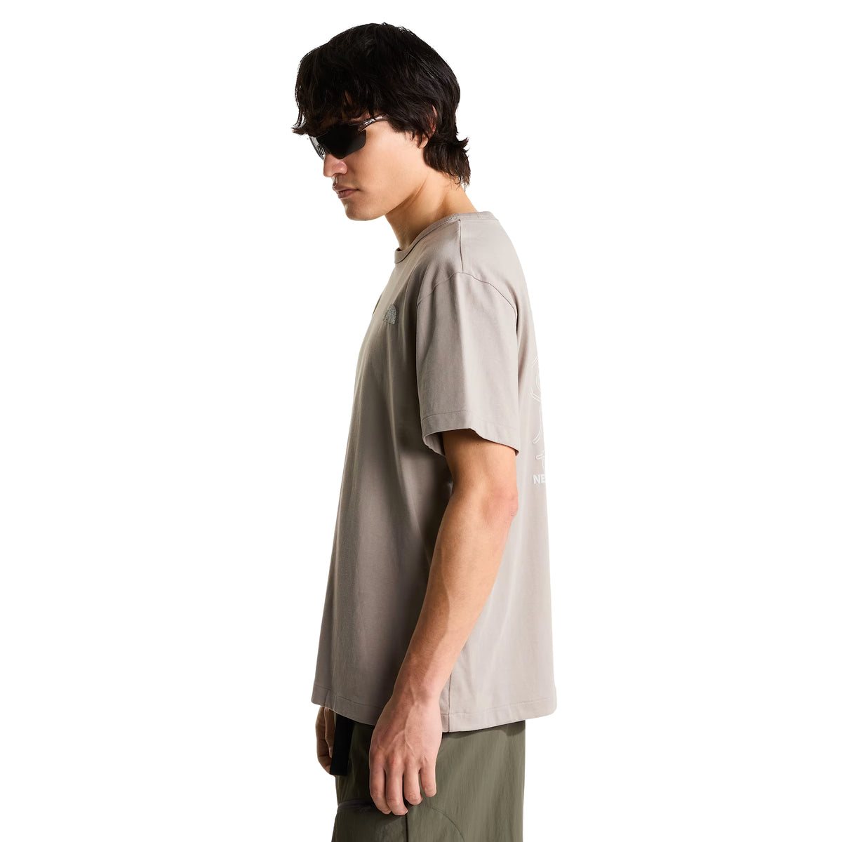 T-shirt The North Face NSE Slopes Relaxed em stone slab — modelo masculino de perfil lateral direito, corpo inteiro, corte relaxed, gráfico nas costas parcialmente visível, calças cargo verde military, óculos de sol escuros rectangulares, fundo branco.