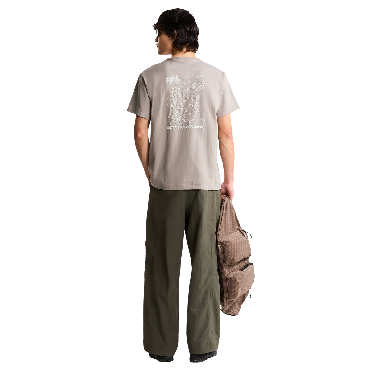 T-shirt The North Face NSE Slopes Relaxed em stone slab — modelo masculino de costas, corpo inteiro, gráfico NSE Slopes com mapa de pistas, "1966" e "NEVER STOP EXPLORING", mochila bege The North Face na mão, calças cargo verde military, fundo branco.