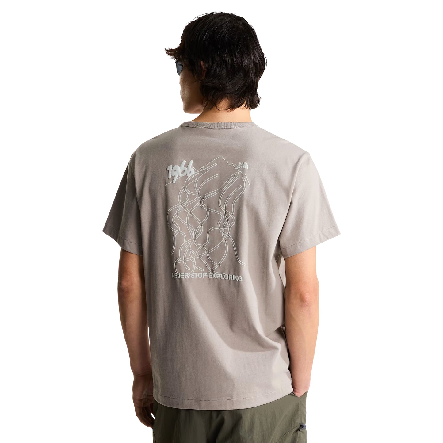 T-shirt The North Face NSE Slopes Relaxed em stone slab — modelo masculino de costas, meio corpo, gráfico NSE Slopes com mapa de pistas em linha, "1966" e logótipo TNF, calças cargo verde military, óculos de sol no decote, fundo branco.
