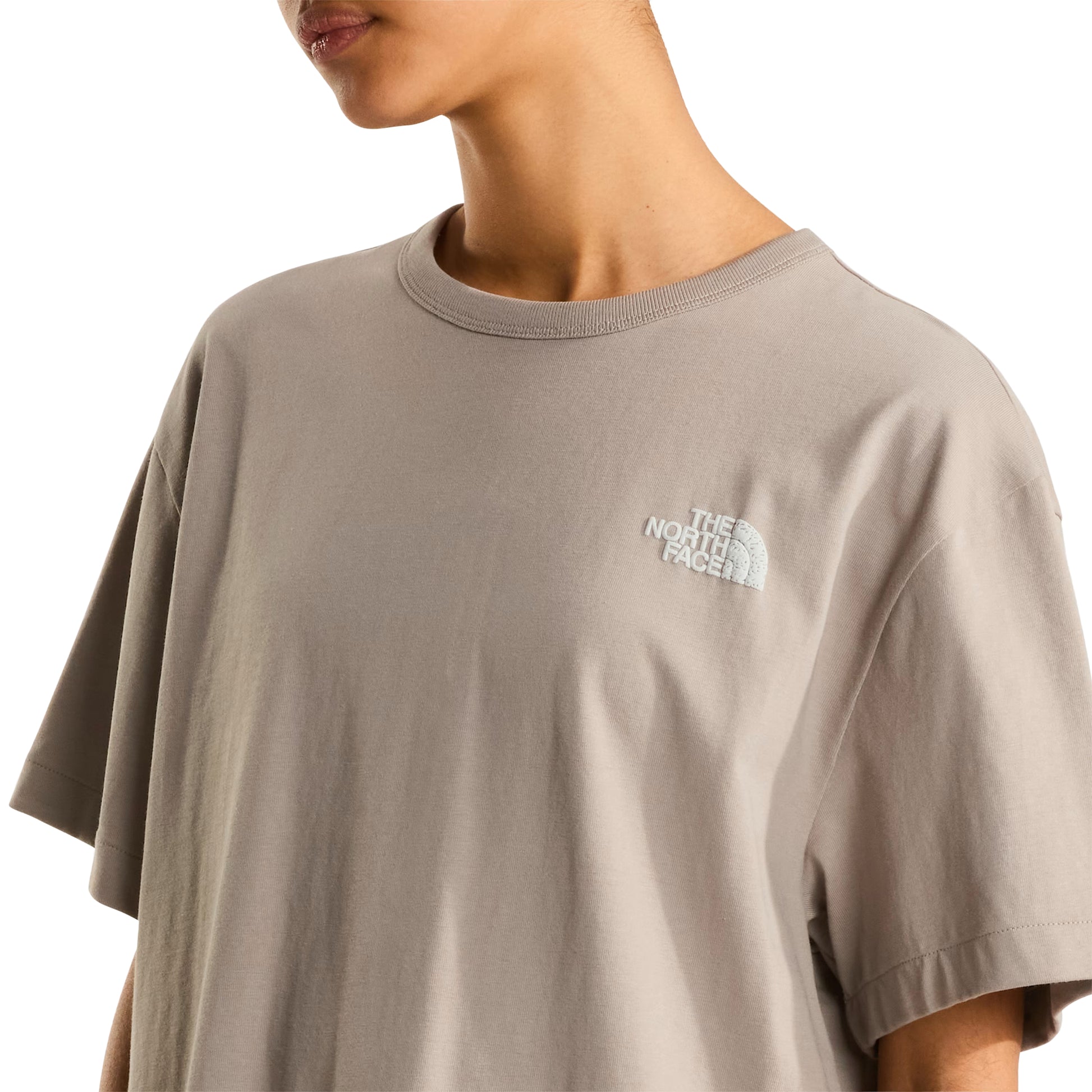 T-shirt The North Face NSE Slopes Relaxed em stone slab — pormenor frontal, meio corpo feminino, logótipo The North Face bordado a branco no peito esquerdo, gola redonda canelada em relevo, tecido jersey stone slab, fundo branco.