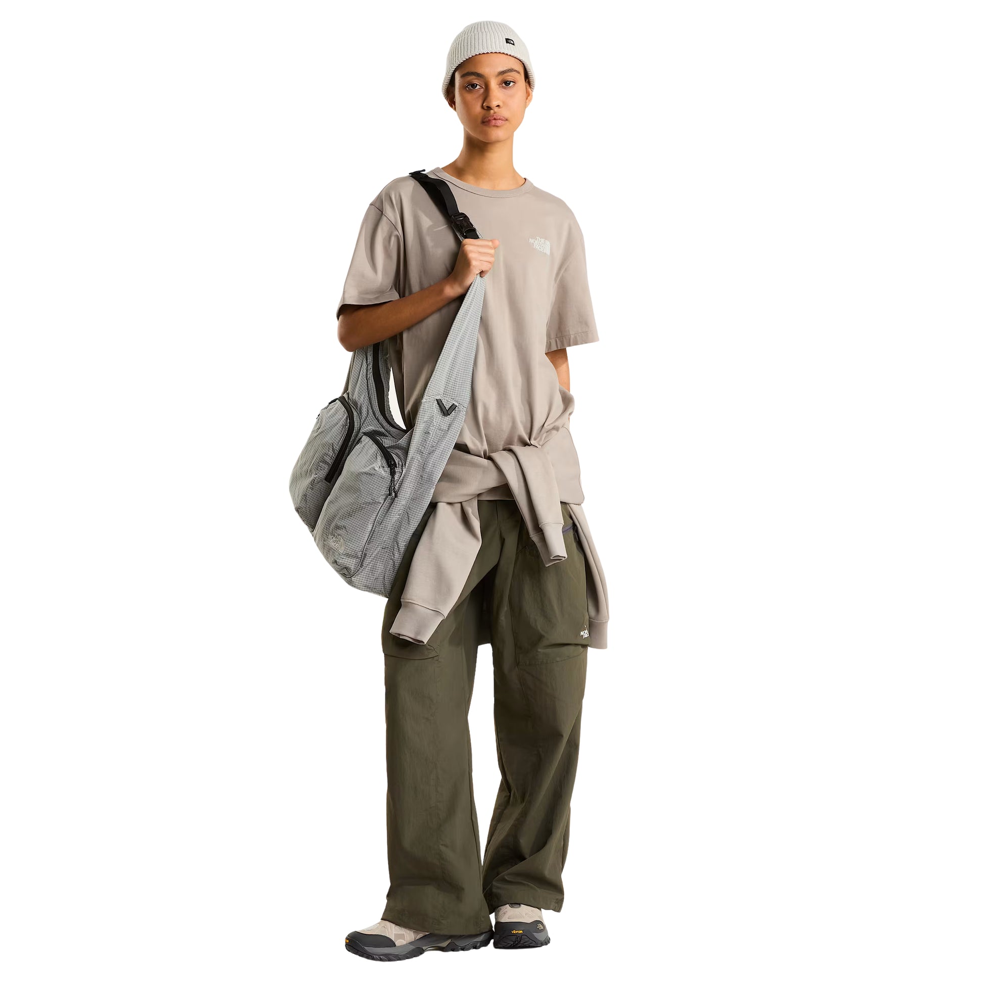 T-shirt The North Face NSE Slopes Relaxed em stone slab — modelo feminino de frente, corpo inteiro, corte relaxed oversized, mala The North Face cinzenta às riscas a tiracolo, gorro canelado branco, calças cargo verde military, sapatilhas cinzentas, fundo branco.