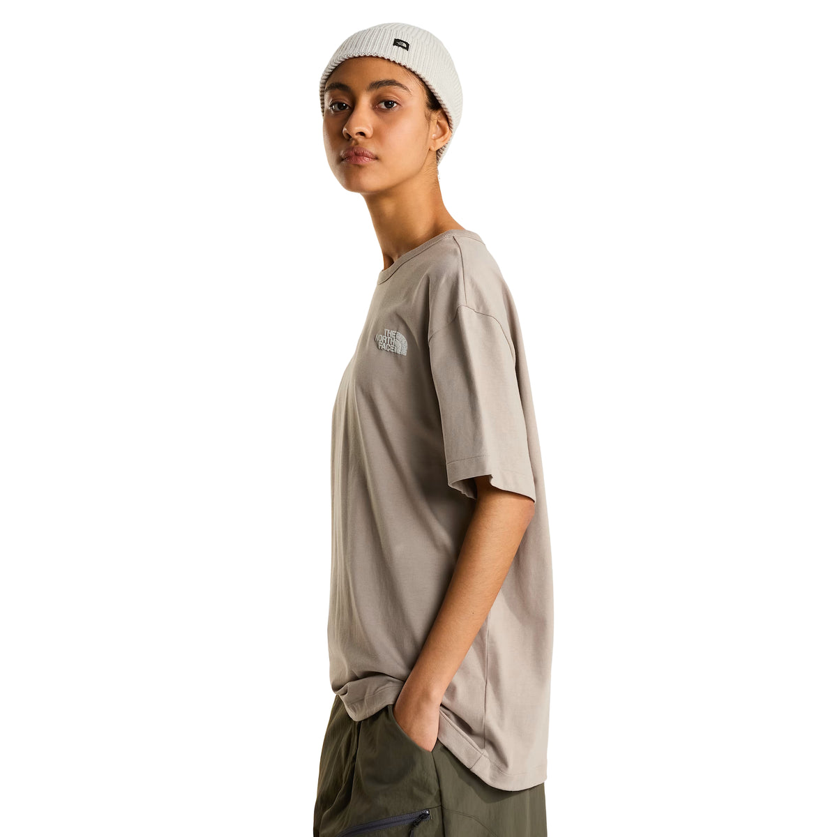 T-shirt The North Face NSE Slopes Relaxed em stone slab — modelo feminino de perfil lateral esquerdo, corpo inteiro, corte relaxed oversized, logótipo The North Face em tom-sobre-tom no peito esquerdo, gola redonda canelada, gorro canelado branco com etiqueta TNF, calças cargo verde military, mão no bolso, fundo branco.