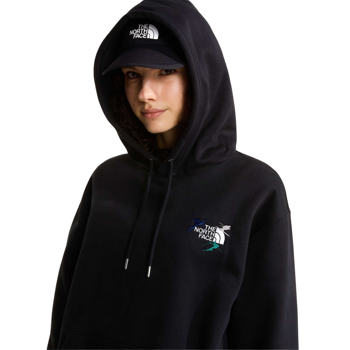 Vista aproximada da sweat The North Face Off Piste Embro preta numa modelo, com capuz colocado sobre o boné e gráficos bordados no peito.